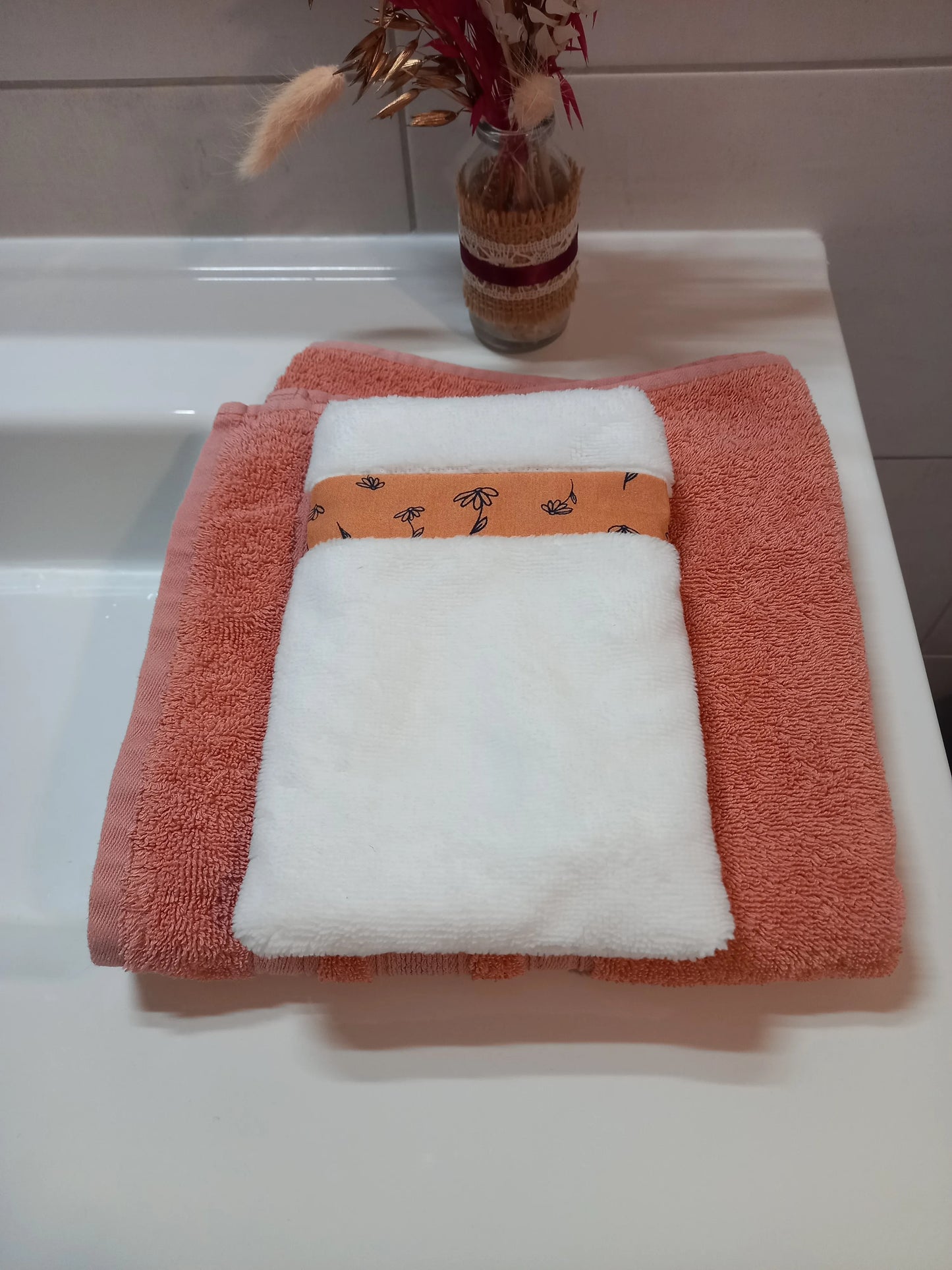 Le gant nettoyant visage de la collection fleurs printanières est posé à plat sur une serviette éponge corail. Il est en tissu éponge blanc avec une bande à motif floral orange.