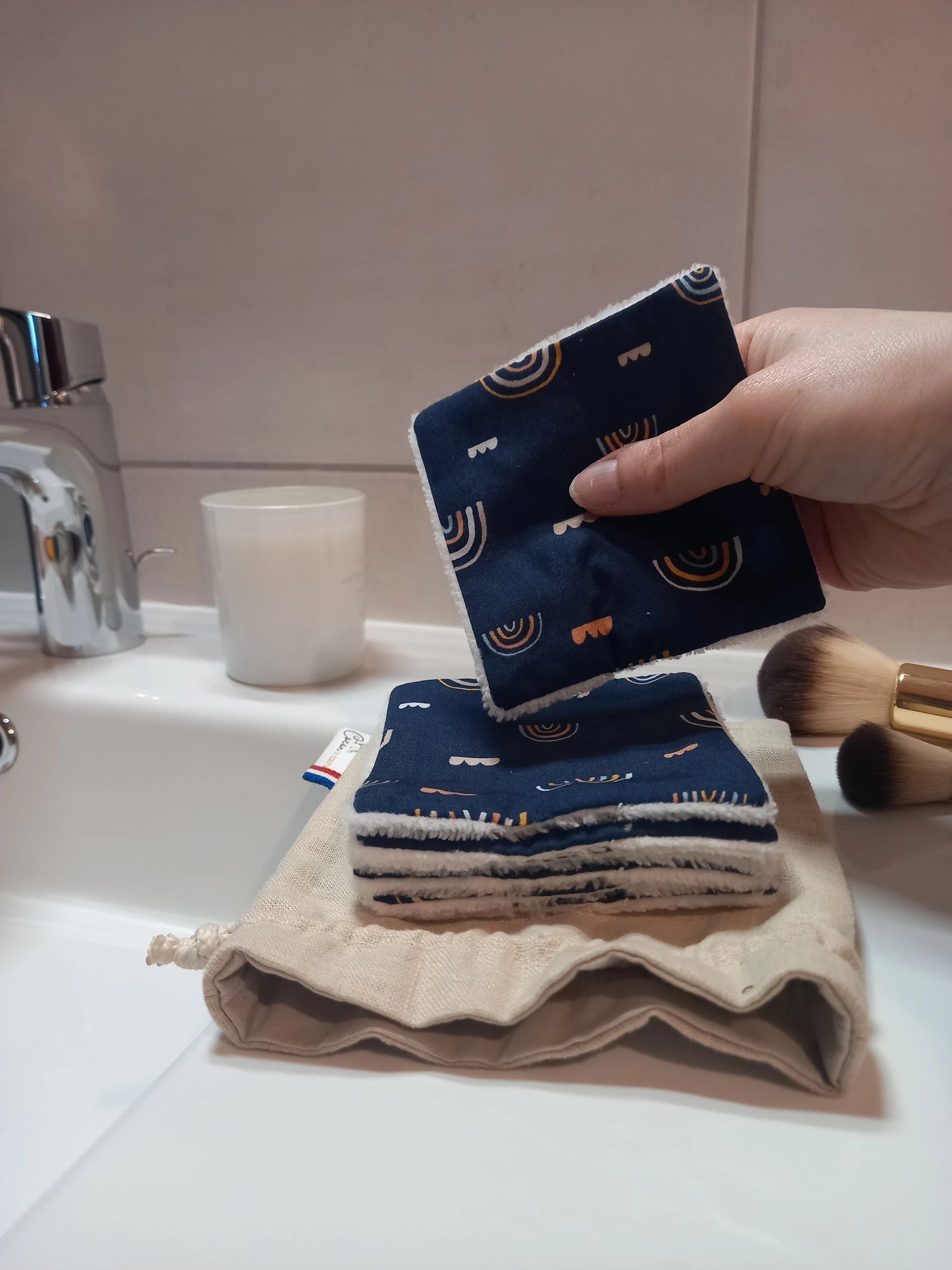 Main tenant un carré démaquillant lavable au motif arcs-en-ciel sur fond bleu marine au-dessus d'une pile de carrés posée sur le pochon en lin, dans une salle de bain avec des pinceaux de maquillage.