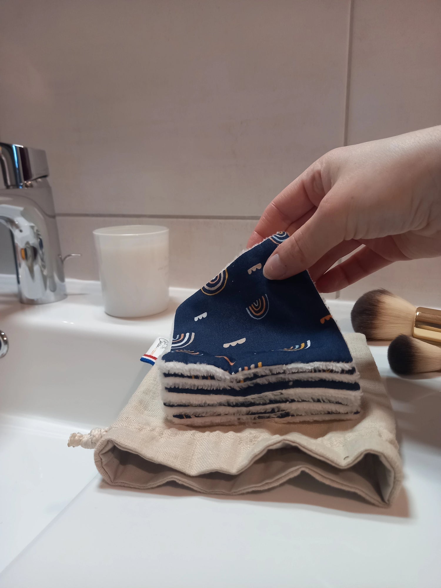 Carré démaquillant lavable au motif arcs-en-ciel sur fond bleu marine tenu en biais entre les doigts, au-dessus d'une pile de carrés posée sur le pochon en lin, sur le rebord d'un lavabo avec des pinceaux de maquillage.