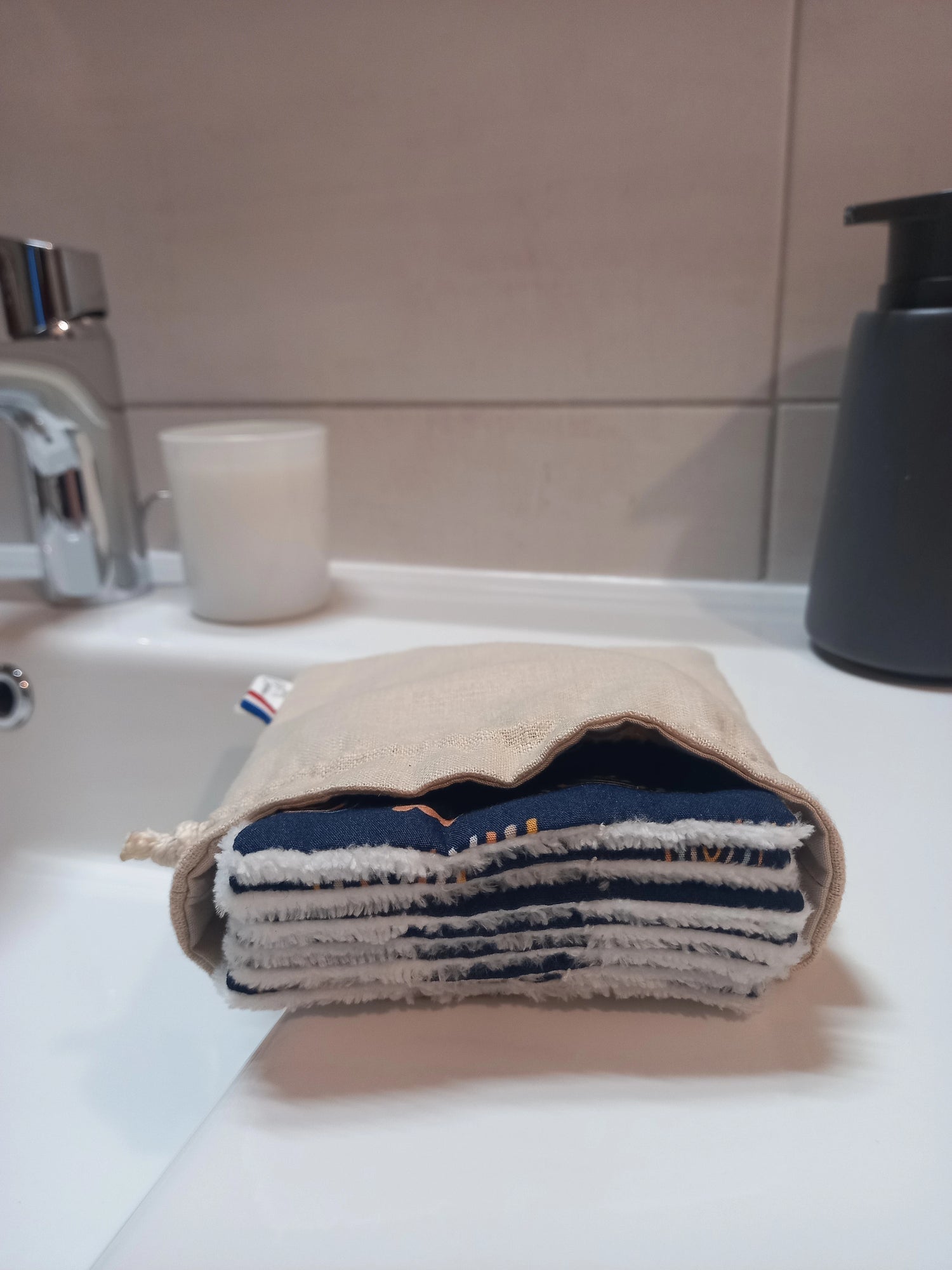 Carrés démaquillants lavables empilés dans leur pochon en lin posé sur le rebord d'un lavabo, le motif arcs-en-ciel sur fond bleu marine et le côté éponge blanc visibles sur la tranche.