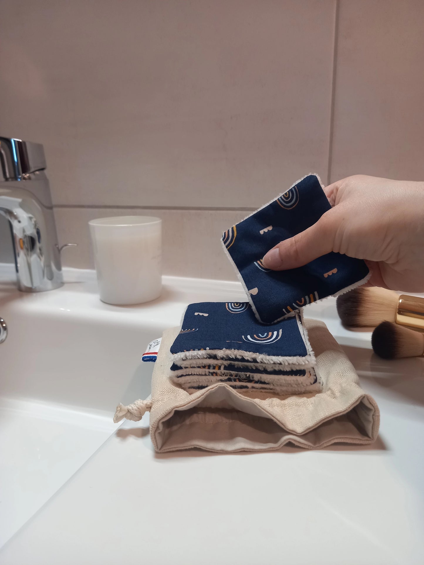 Carré démaquillant lavable au motif arcs-en-ciel sur fond bleu marine tenu en biais entre les doigts, au-dessus de la pile posée sur le pochon en lin, sur le rebord d'un lavabo avec des pinceaux de maquillage.
