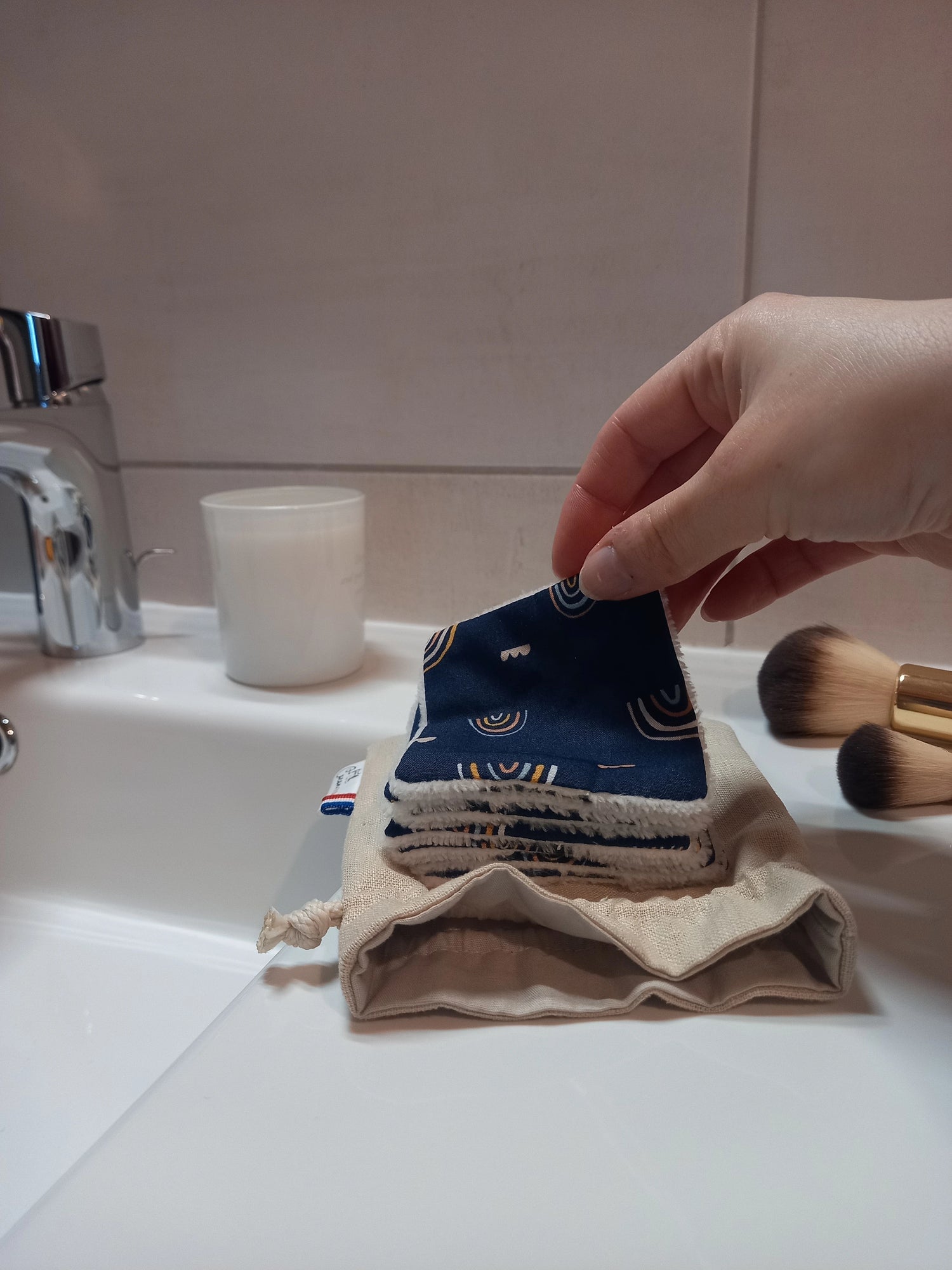 Main tenant un carré démaquillant lavable au motif arcs-en-ciel sur fond bleu marine au-dessus d'une pile de carrés posée sur le pochon en lin ouvert, dans une salle de bain avec des pinceaux de maquillage.