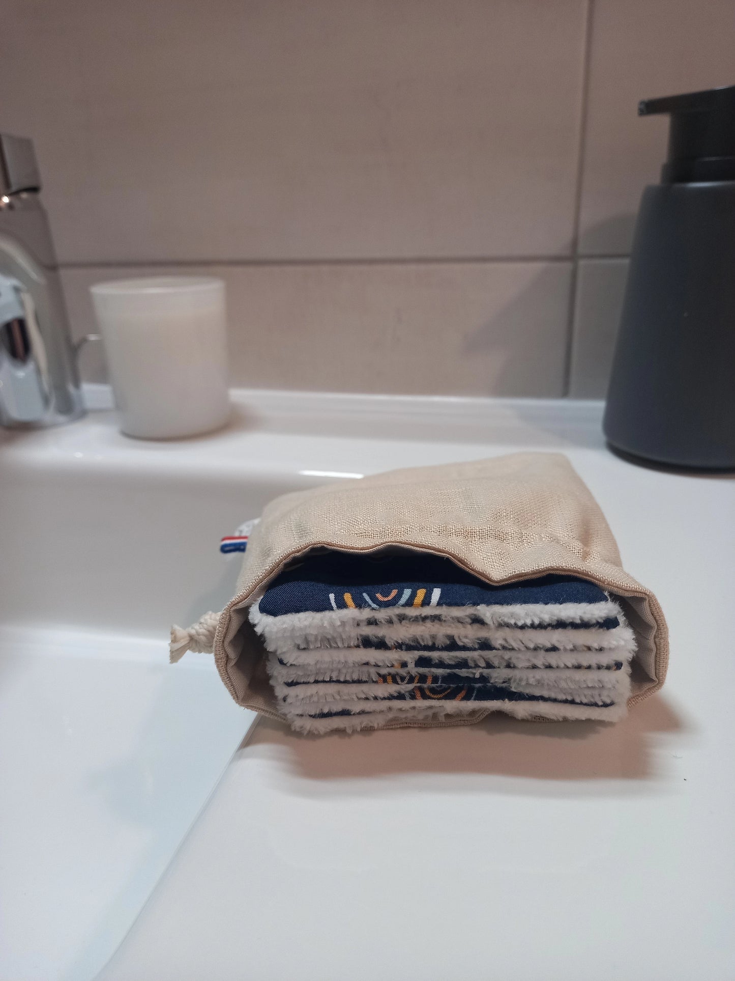 Carrés démaquillants lavables empilés dans leur pochon en lin posé sur le rebord d'un lavabo, le motif arcs-en-ciel sur fond bleu marine et le côté éponge blanc visibles sur la tranche.
