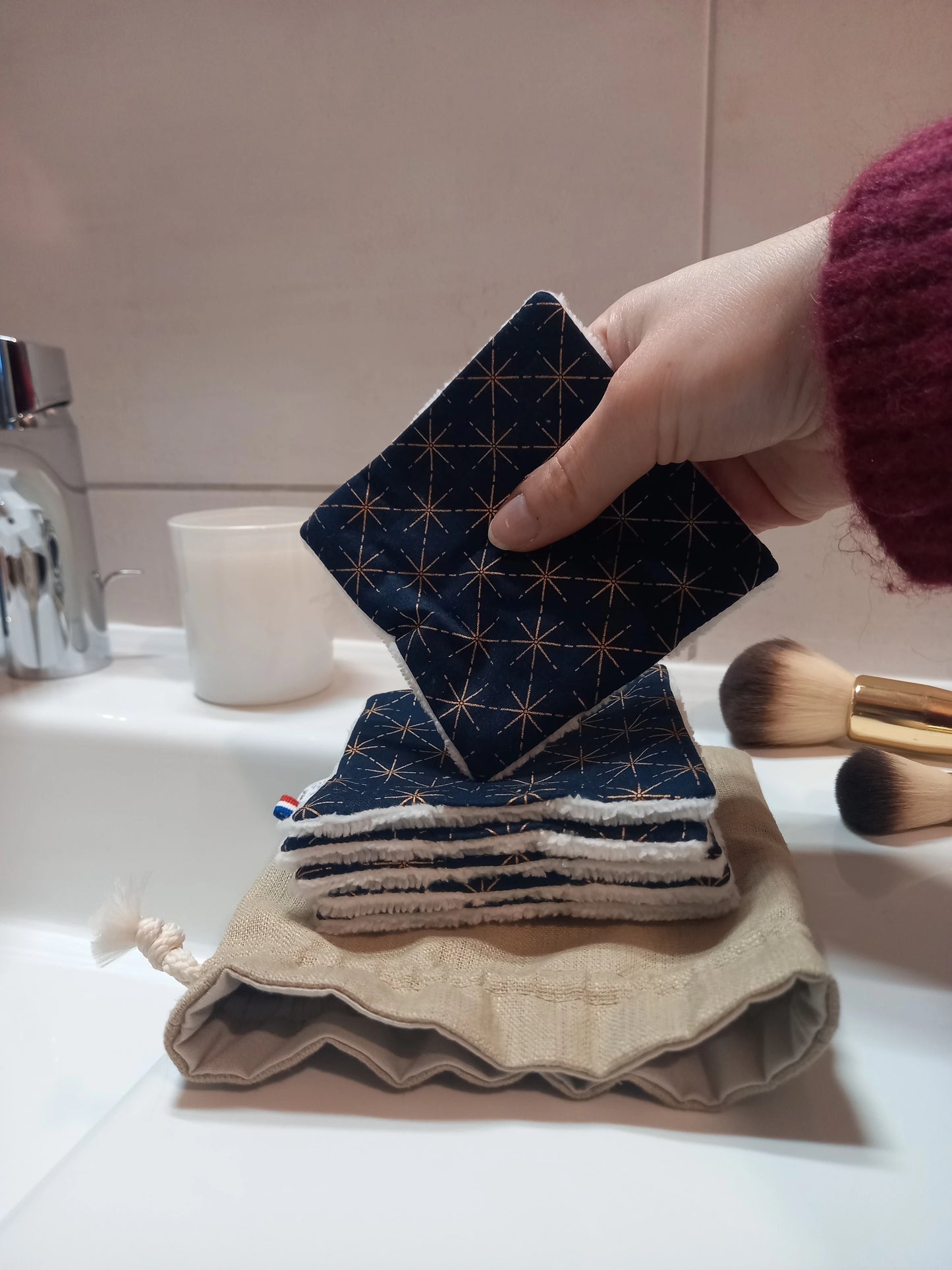 Main tenant un carré démaquillant lavable au motif étoiles dorées sur fond bleu marine en biais au-dessus d'une pile de carrés posée sur le pochon en lin, dans une salle de bain avec des pinceaux de maquillage.
