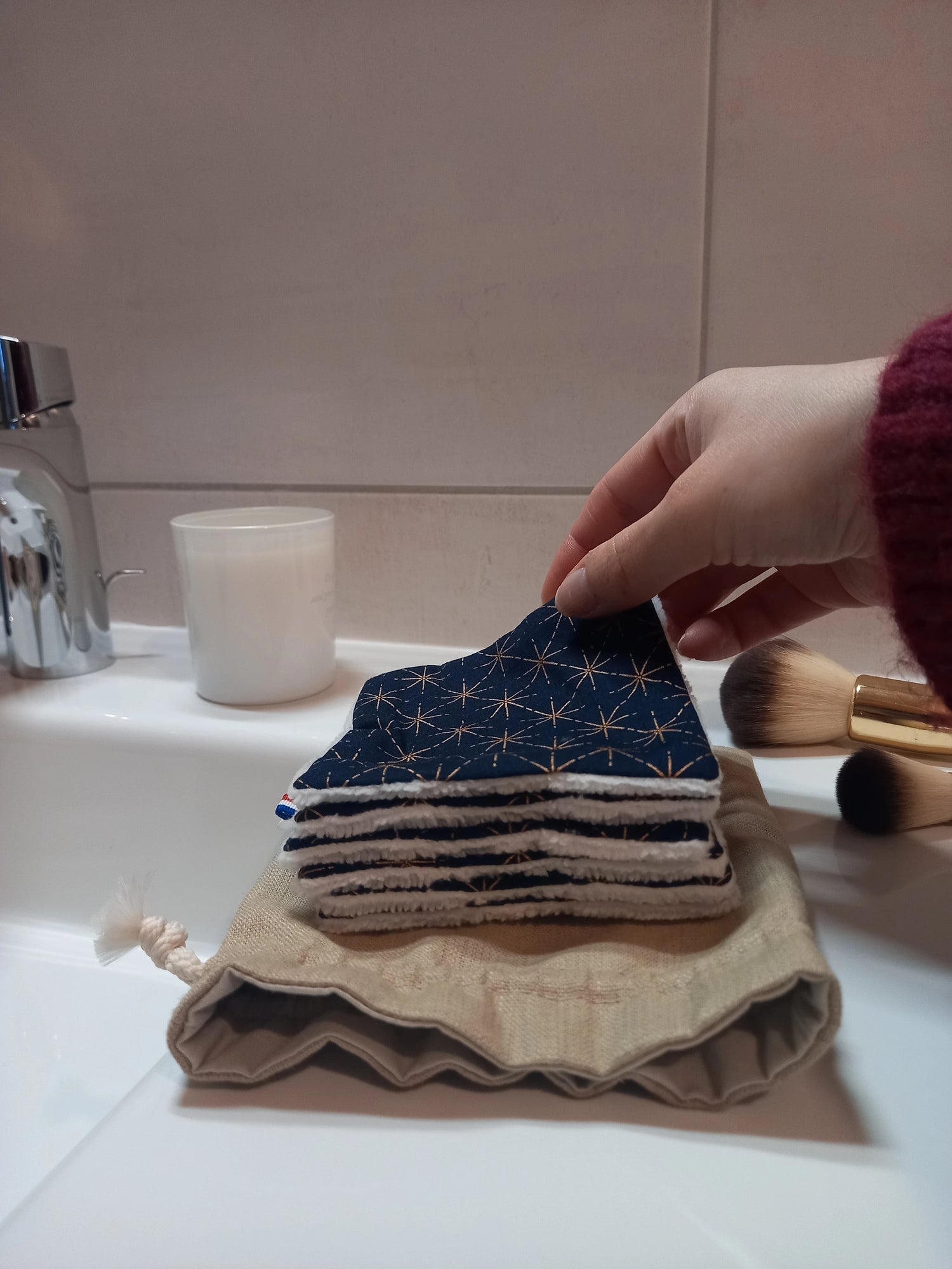 Carré démaquillant lavable au motif étoiles dorées sur fond bleu marine tenu entre les doigts, au-dessus d'une pile de carrés posée sur le pochon en lin, sur le rebord d'un lavabo avec des pinceaux de maquillage.