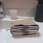 Carrés démaquillants lavables empilés dans leur pochon en lin posé sur le rebord d'un lavabo, le motif étoiles dorées sur fond bleu marine et le côté éponge blanc visibles sur la tranche.