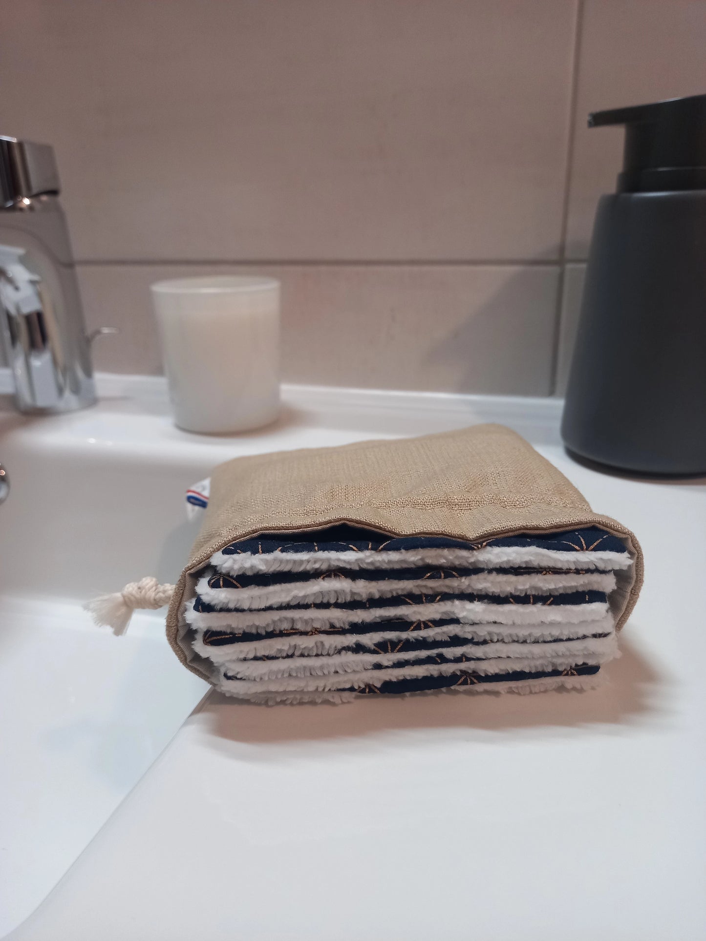 Carrés démaquillants lavables empilés dans leur pochon en lin posé sur le rebord d'un lavabo, le motif étoiles dorées sur fond bleu marine et le côté éponge blanc visibles sur la tranche.