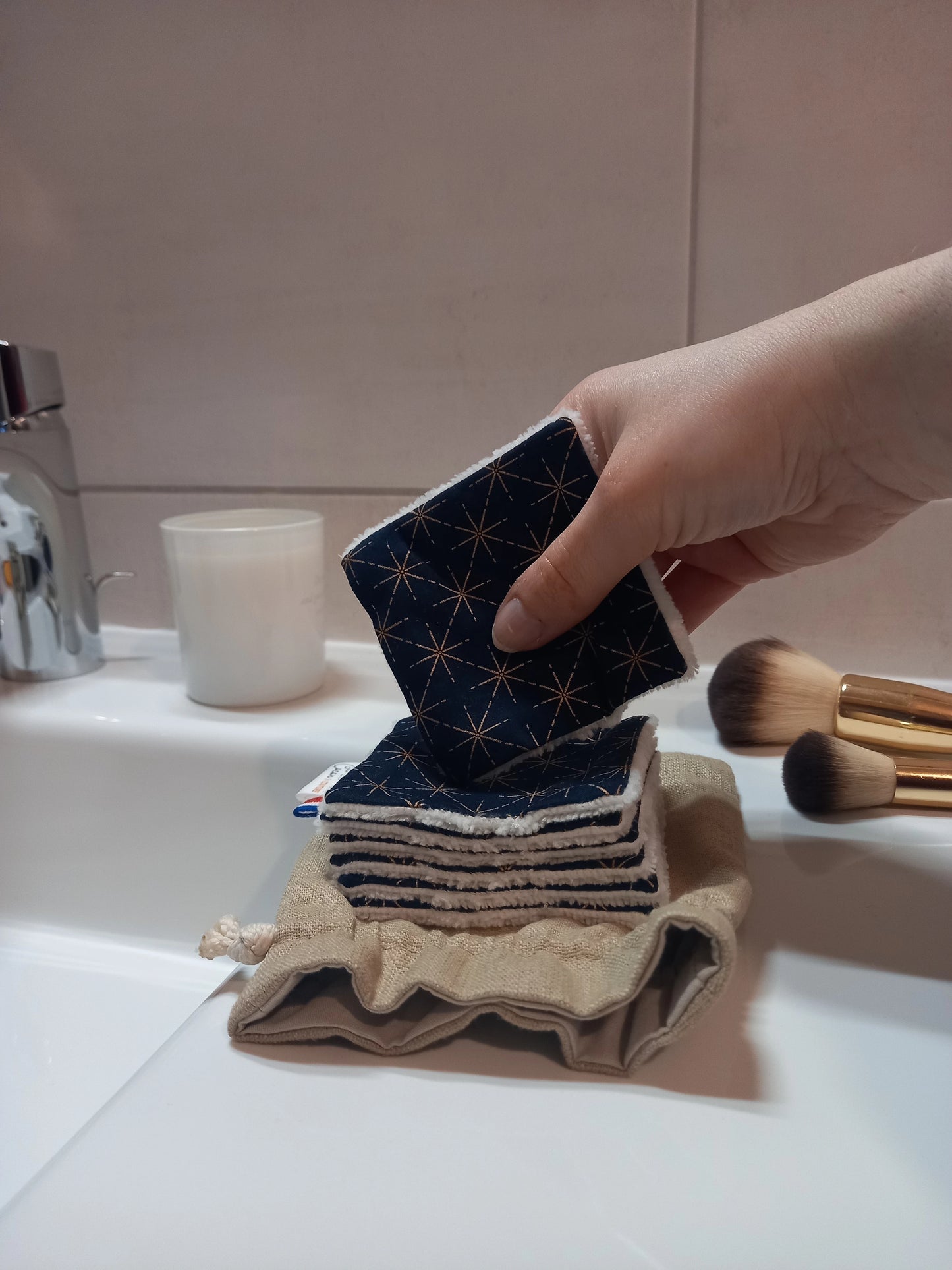 Main tenant un carré démaquillant lavable au motif étoiles dorées sur fond bleu marine en biais au-dessus d'une pile de carrés posée sur le pochon en lin, dans une salle de bain avec des pinceaux de maquillage.