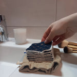 Carré démaquillant lavable au motif étoiles dorées sur fond bleu marine tenu entre les doigts, au-dessus d'une pile de carrés posée sur le pochon en lin, sur le rebord d'un lavabo avec des pinceaux de maquillage.