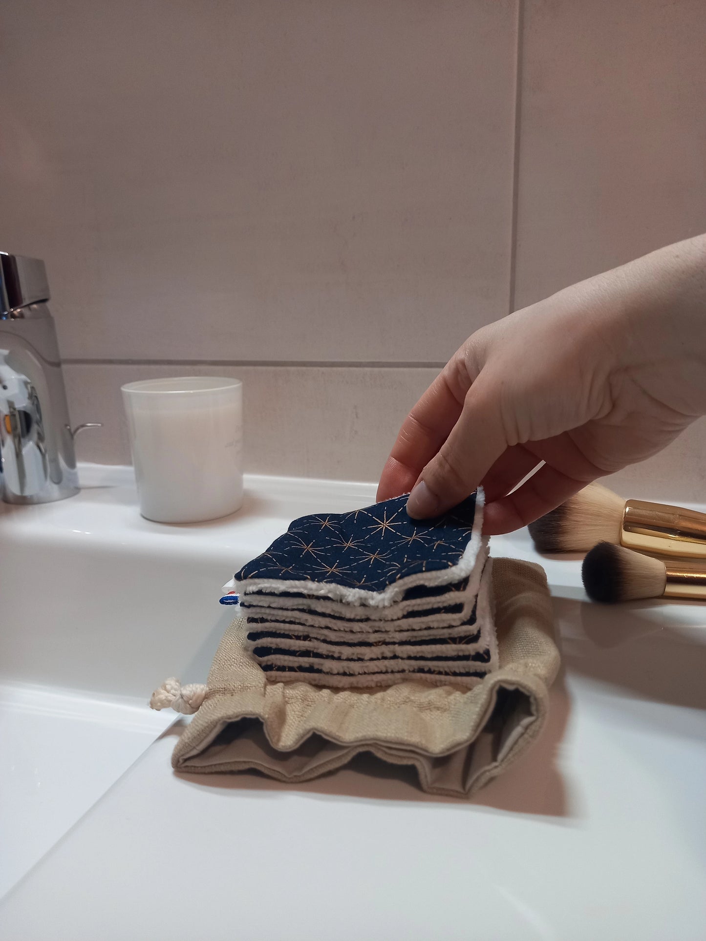 Carré démaquillant lavable au motif étoiles dorées sur fond bleu marine tenu entre les doigts, au-dessus d'une pile de carrés posée sur le pochon en lin, sur le rebord d'un lavabo avec des pinceaux de maquillage.