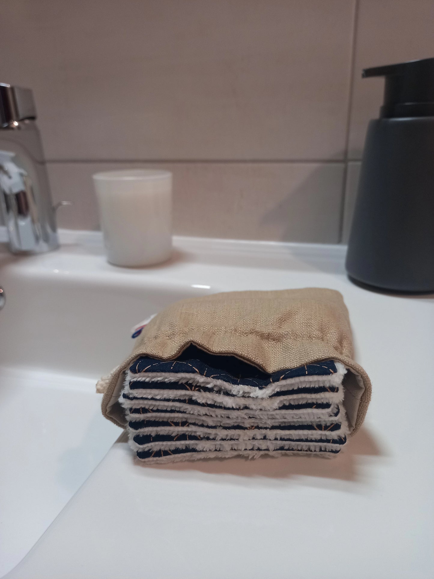 Carrés démaquillants lavables empilés dans leur pochon en lin posé sur le rebord d'un lavabo, le motif étoiles dorées sur fond bleu marine et le côté éponge blanc visibles sur la tranche.