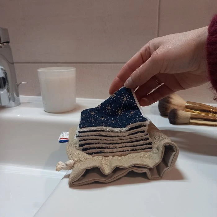 Main qui prend un carré démaquillant lavable Ciel Étoilé au-dessus de son pochon en lin ouvert, des pinceaux de maquillage sont visibles en arrière plan