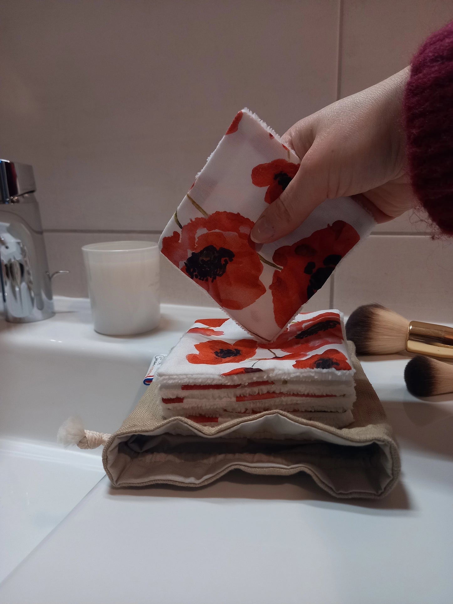 Main tenant un carré démaquillant lavable au motif coquelicots rouges sur fond blanc au-dessus d'une pile de carrés posée sur le pochon en lin, dans une salle de bain avec des pinceaux de maquillage.