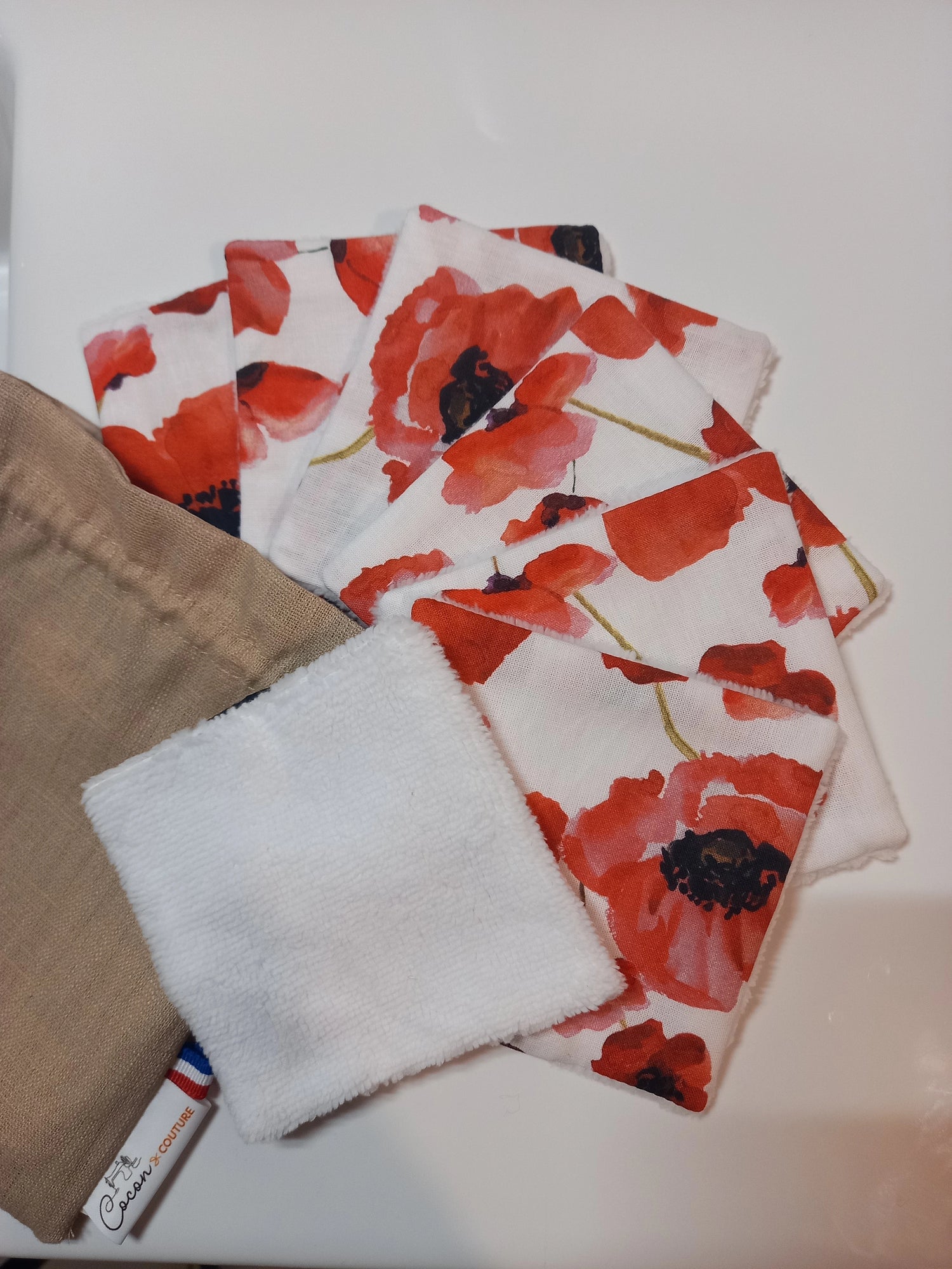Lot de sept carrés démaquillants lavables au motif coquelicots rouges sur fond blanc, disposés en éventail à côté de leur pochon en lin beige Cocon Couture, un carré retourné montrant le côté éponge blanc.