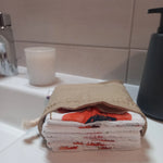 Carrés démaquillants lavables empilés dans leur pochon en lin posé sur le rebord d'un lavabo, l'empilement laissant voir le motif coquelicots rouges et le côté éponge blanc sur la tranche.