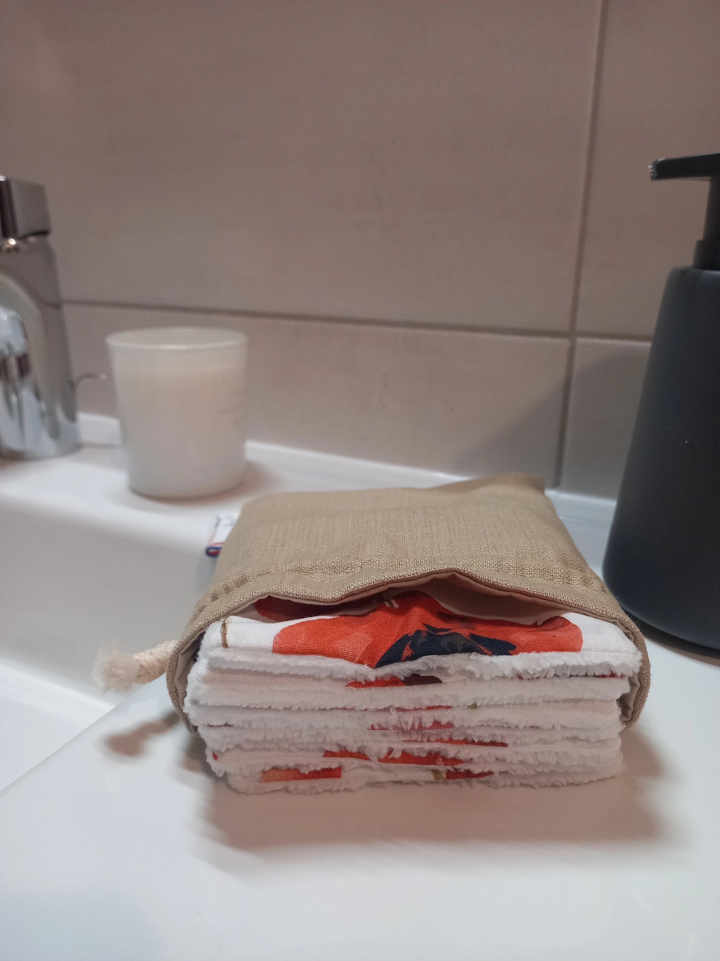 Carrés démaquillants lavables empilés dans leur pochon en lin posé sur le rebord d'un lavabo, l'empilement laissant voir le motif coquelicots rouges et le côté éponge blanc sur la tranche.