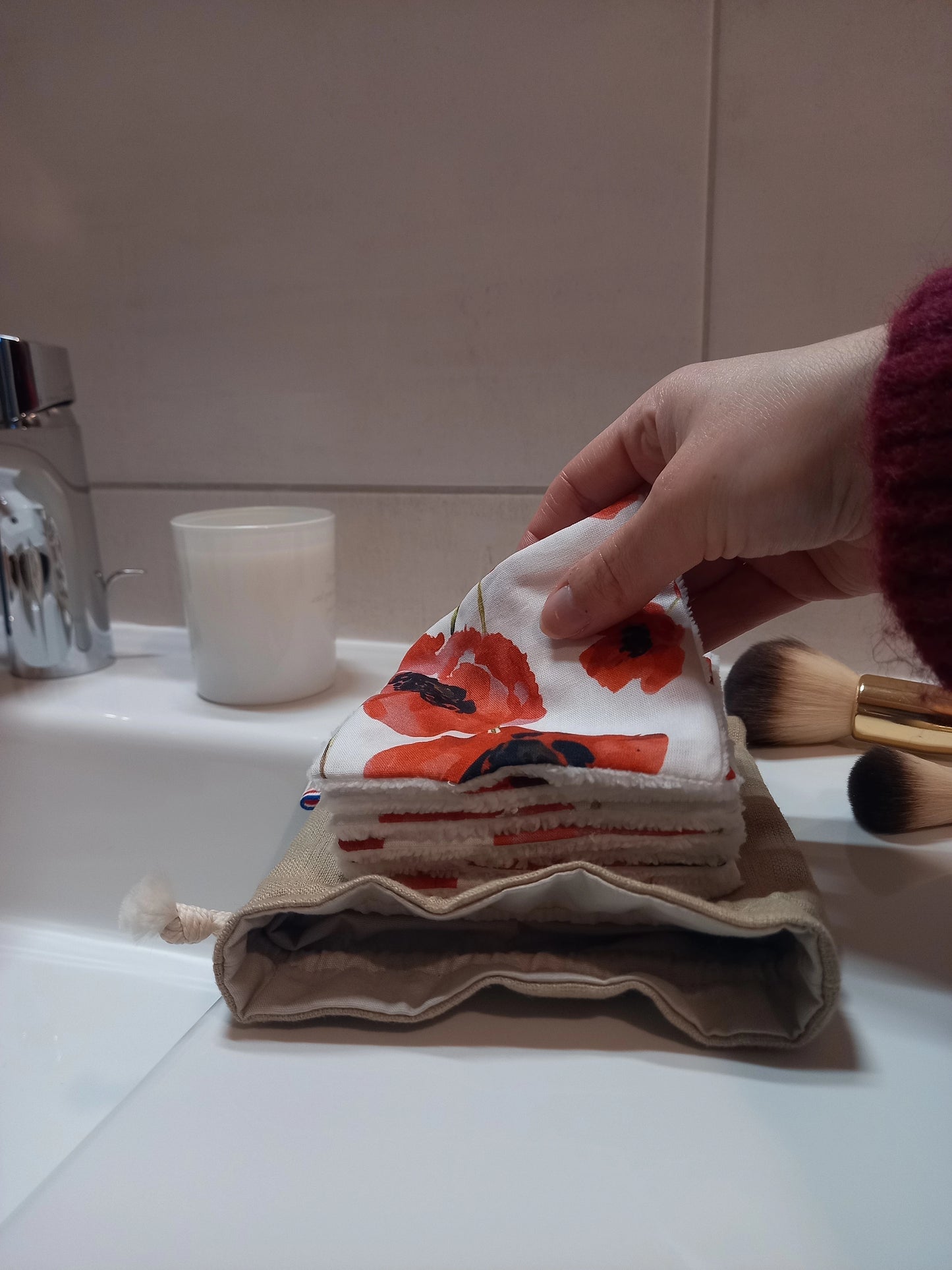 Carré démaquillant lavable au motif coquelicots rouges sur fond blanc tenu entre les doigts, au-dessus d'une pile de carrés posée sur le pochon en lin, sur le rebord d'un lavabo avec un pinceau de maquillage à côté.