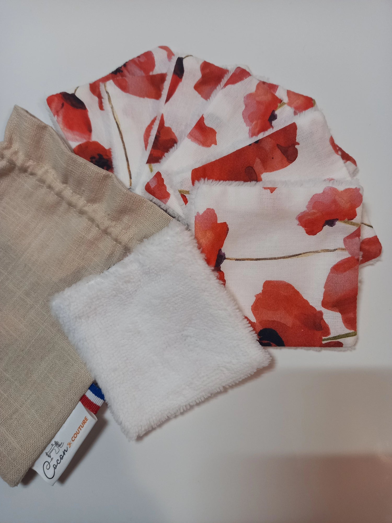 Carrés démaquillants lavables en éventail, le carré de devant retourné sur son côté éponge blanc, les autres laissant voir le motif coquelicots rouges, à côté du pochon en lin Cocon Couture.
