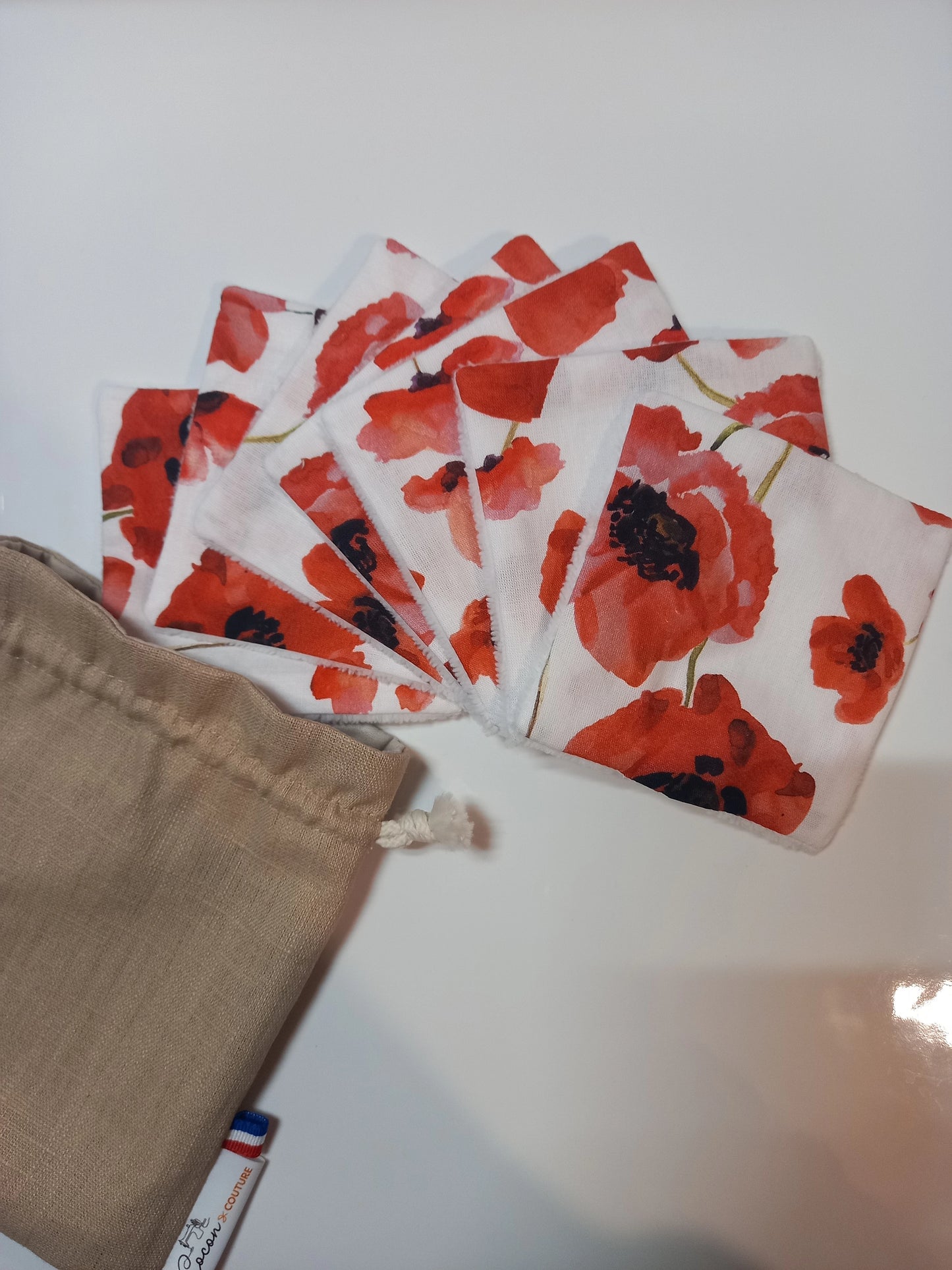 Lot de sept carrés démaquillants lavables au motif coquelicots rouges sur fond blanc, disposés en éventail à côté de leur pochon en lin beige Cocon Couture.