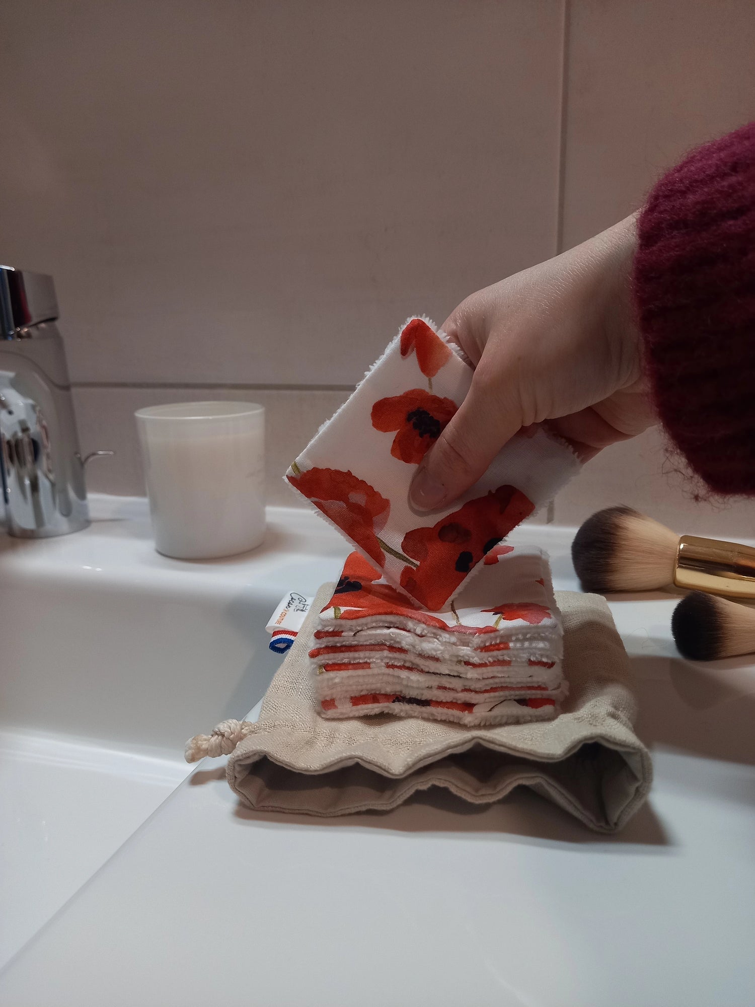 Carré démaquillant lavable au motif coquelicots rouges tenu en biais entre les doigts, au-dessus de la pile posée sur le pochon en lin, sur le rebord d'un lavabo avec des pinceaux de maquillage.
