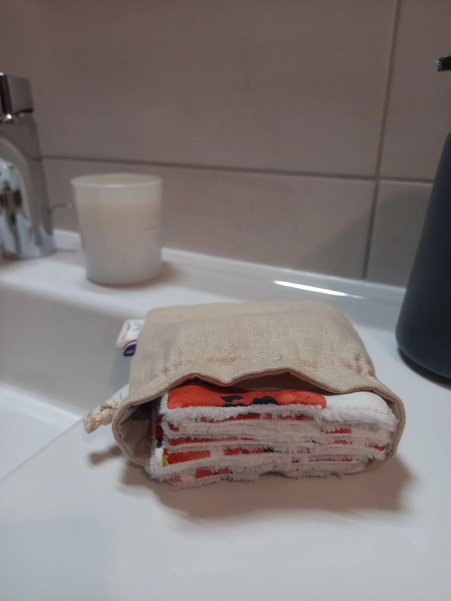 Carrés démaquillants lavables empilés dans leur pochon en lin sur le rebord d'un lavabo, l'empilement laissant voir l'alternance du motif coquelicots rouges et du côté éponge blanc.