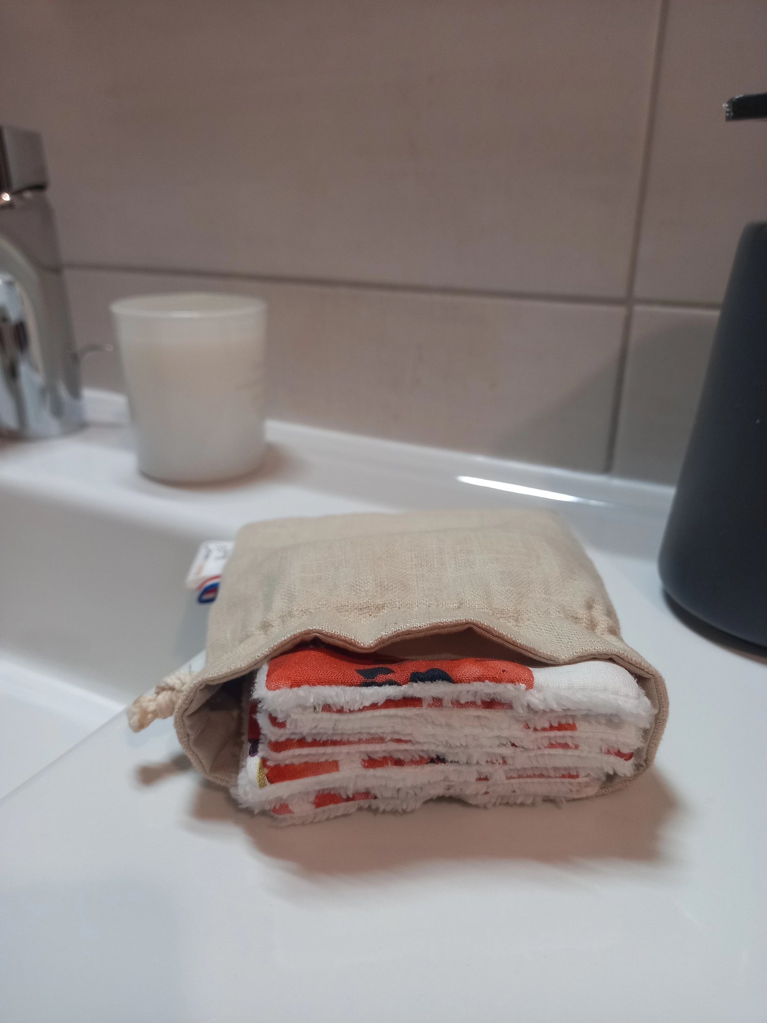 Carrés démaquillants lavables empilés dans leur pochon en lin posé sur le rebord d'un lavabo, le motif coquelicots rouges et le côté éponge blanc visibles sur la tranche.