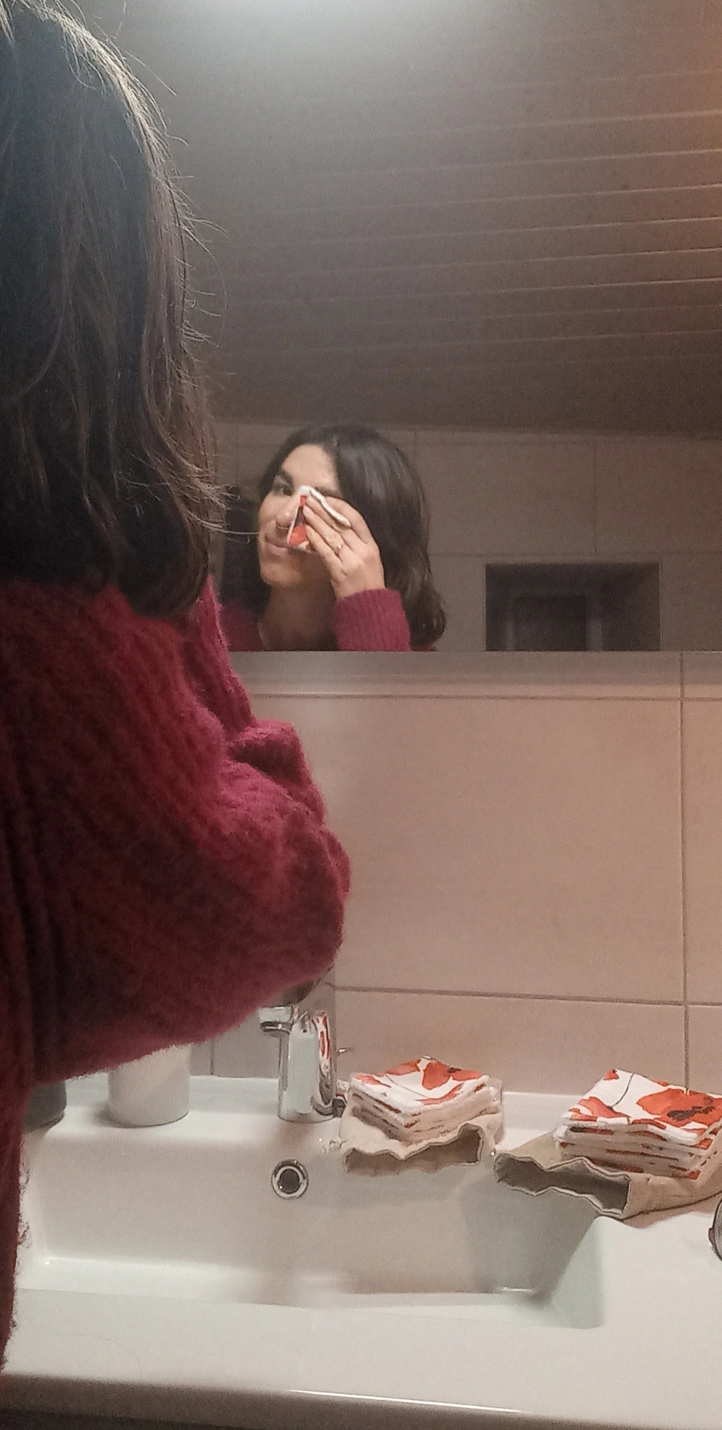 Femme se démaquillant le visage avec un carré démaquillant lavable Coquelicots devant son miroir de salle de bain.