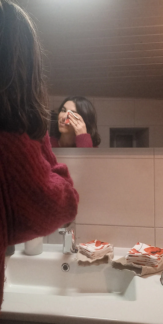 Femme se démaquillant le visage avec un carré démaquillant lavable Coquelicots devant son miroir de salle de bain.