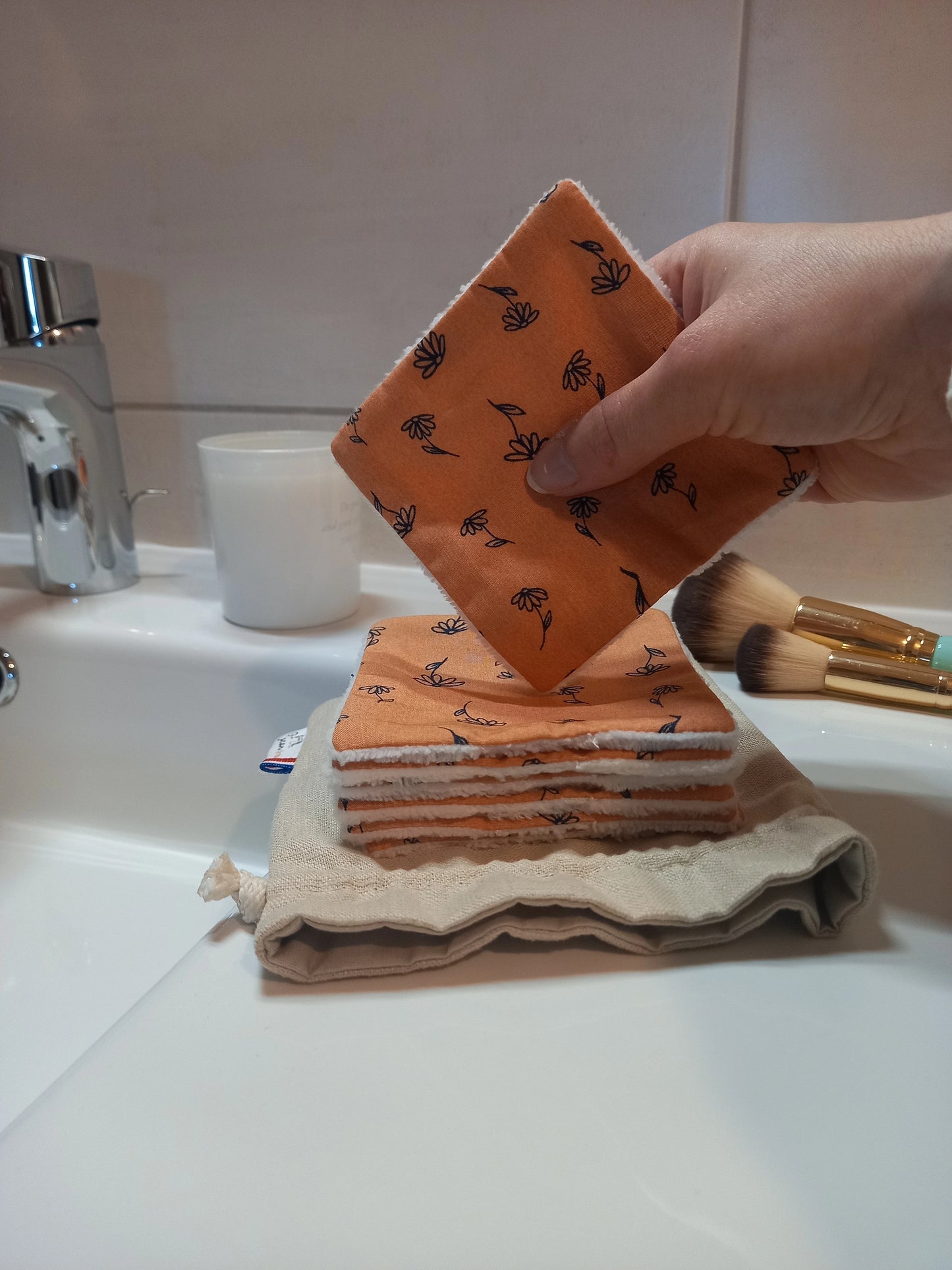 Main tenant un carré démaquillant lavable au motif fleurs printanières orange au-dessus d'une pile de carrés posée sur le pochon en lin, sur un plan de lavabo avec des pinceaux de maquillage à côté.