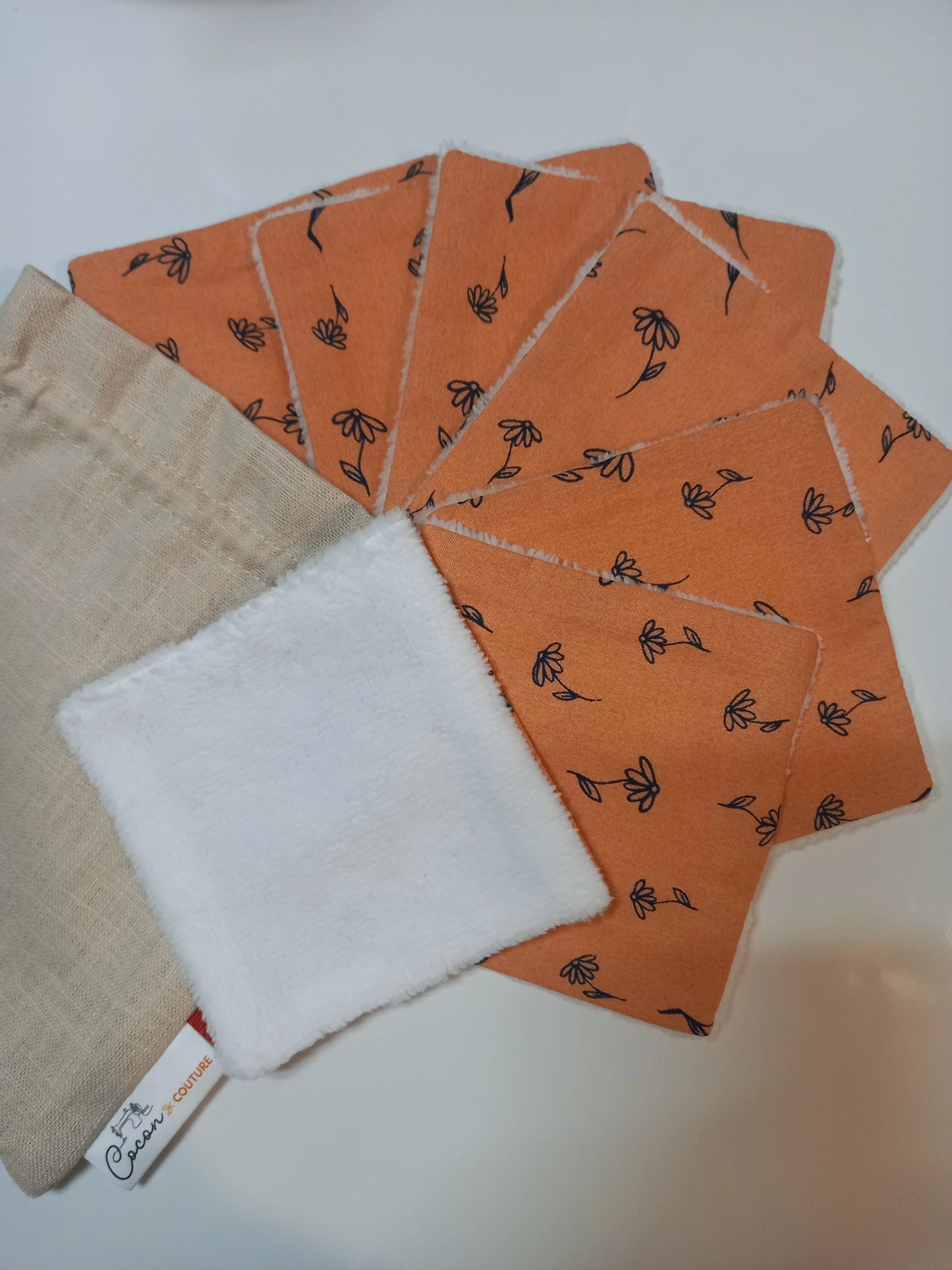 Lot de sept carrés démaquillants lavables au motif fleurs printanières orange, disposés en éventail sur fond blanc à côté de leur pochon en lin beige Cocon Couture, un carré retourné montrant le côté éponge blanc.