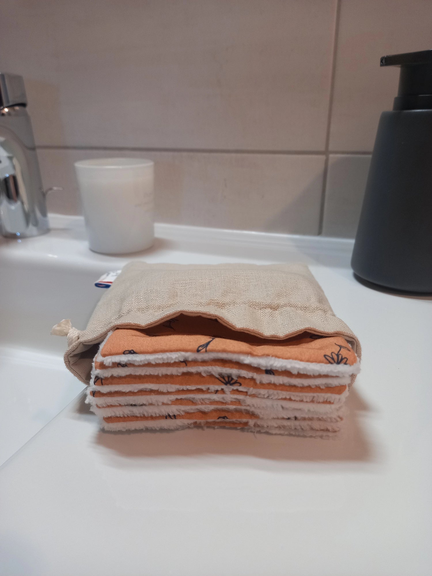 Carrés démaquillants lavables empilés dans leur pochon en lin ouvert, posés sur le rebord d'un lavabo, le côté orange fleuri et le côté éponge blanc visibles sur la tranche.