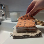 Carré démaquillant lavable tenu entre les doigts, côté orange fleuri face à l'objectif, au-dessus de la pile et de son pochon en lin posés sur le rebord du lavabo.