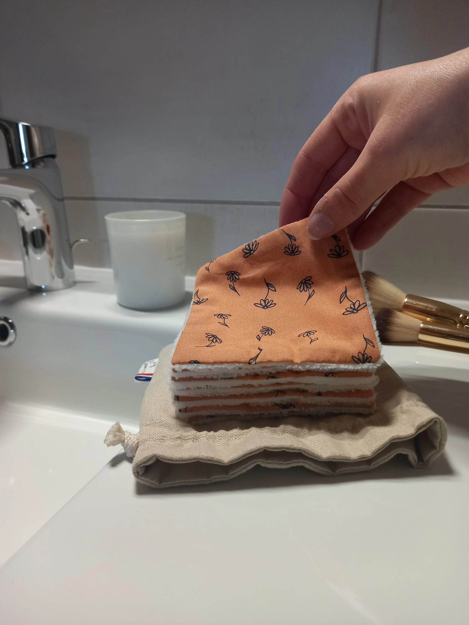 Carré démaquillant lavable tenu entre les doigts, côté orange fleuri face à l'objectif, au-dessus de la pile et de son pochon en lin posés sur le rebord du lavabo.