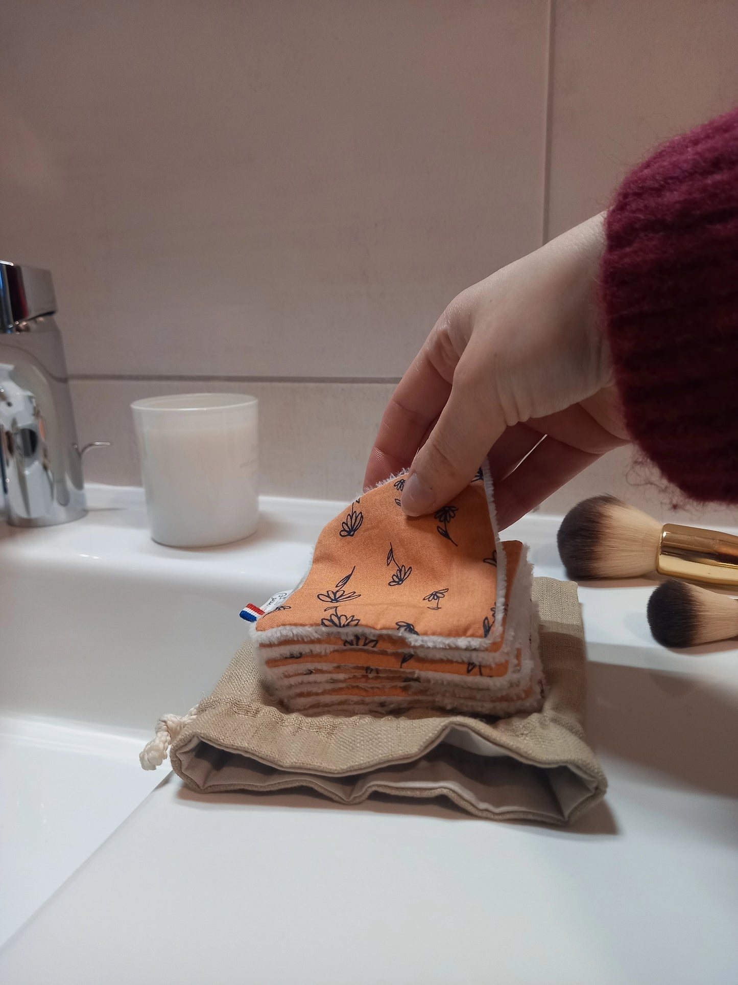Carré démaquillant lavable tenu à la main, côté éponge en bambou blanc apparent, posé sur une pile et son pochon en lin dans une salle de bain.