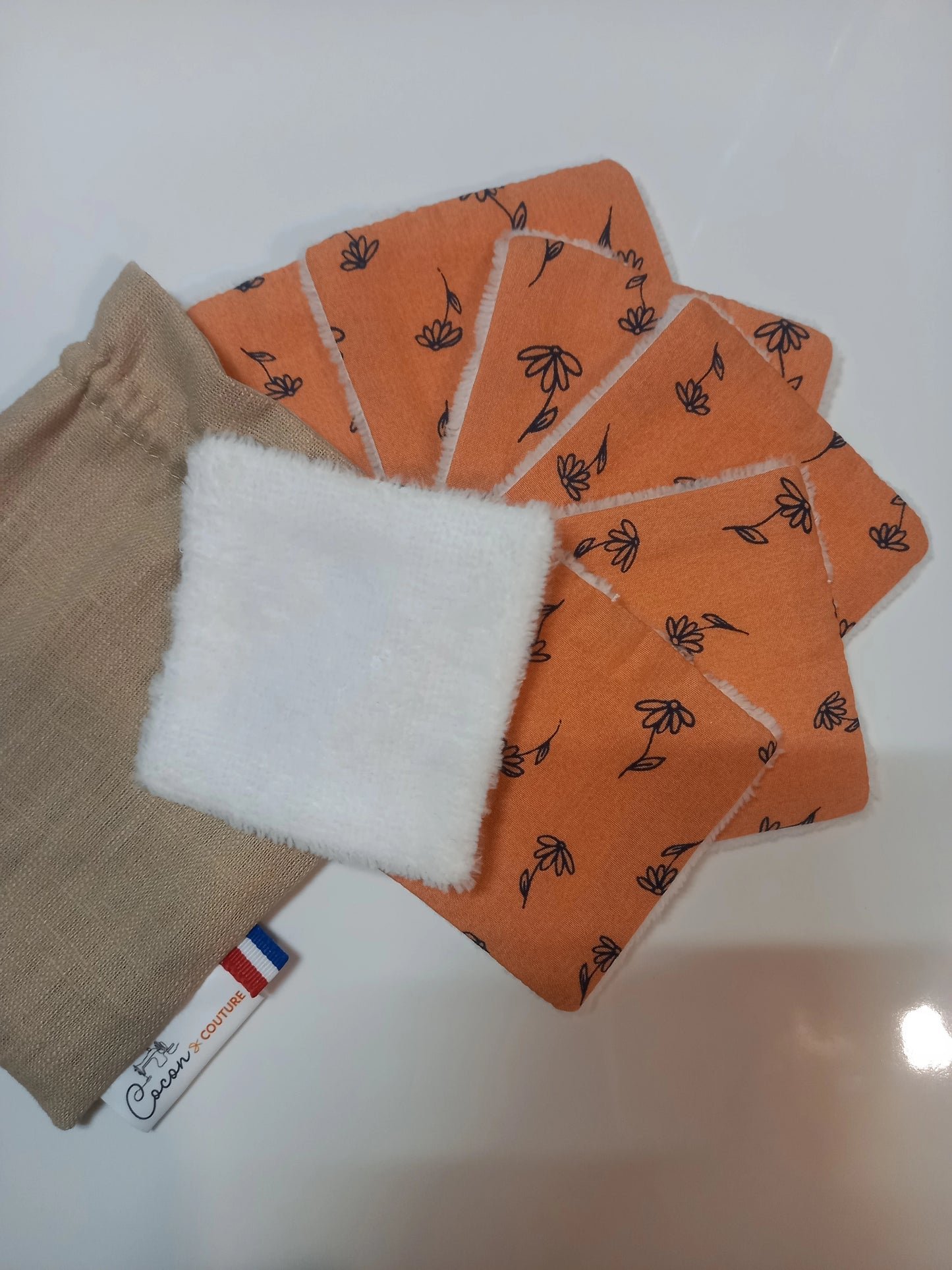 Lot de sept carrés démaquillants lavables Cocon Couture au motif fleurs printanières orange, disposés en éventail avec un carré retourné sur le côté éponge blanc, à côté de leur pochon en lin beige.