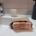 Carrés démaquillants lavables empilés dans leur pochon en lin posé sur un rebord de lavabo, le motif fleurs printanières orange et le côté éponge blanc bien visibles.
