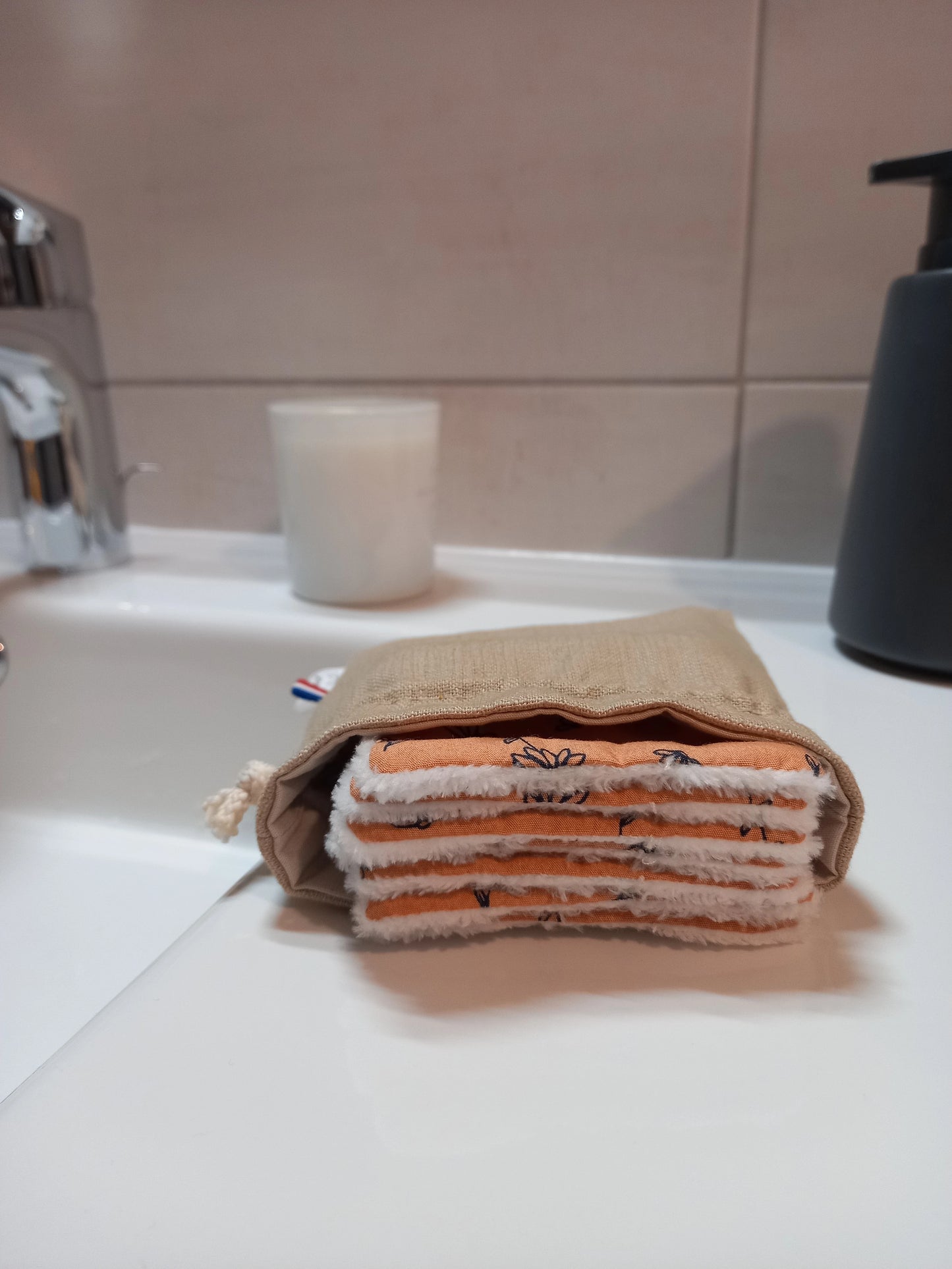 Carrés démaquillants lavables empilés dans leur pochon en lin posé sur un rebord de lavabo, le motif fleurs printanières orange et le côté éponge blanc bien visibles.
