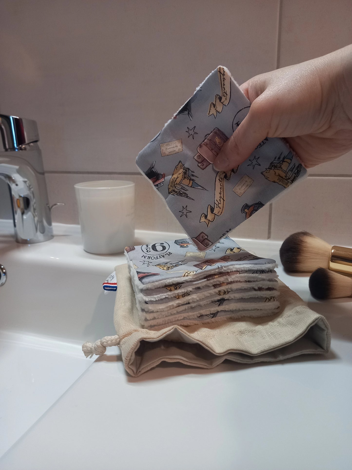Main tenant un carré démaquillant lavable au motif magique gris à train, château et valises au-dessus d'une pile de carrés posée sur le pochon en lin, dans une salle de bain avec des pinceaux de maquillage.