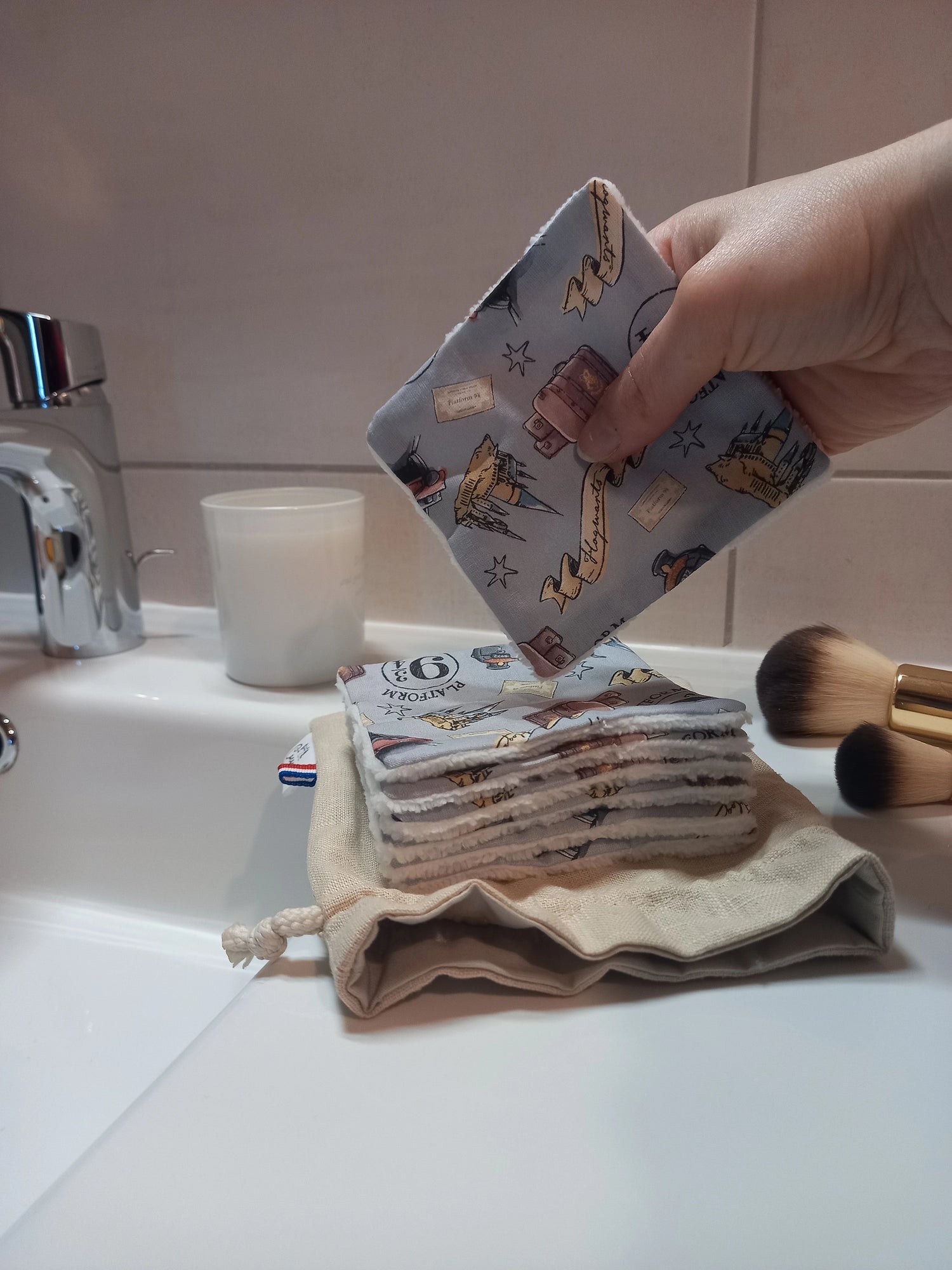 Main tenant un carré démaquillant lavable au motif magique gris à train, château et valises au-dessus d'une pile de carrés posée sur le pochon en lin, dans une salle de bain avec des pinceaux de maquillage.