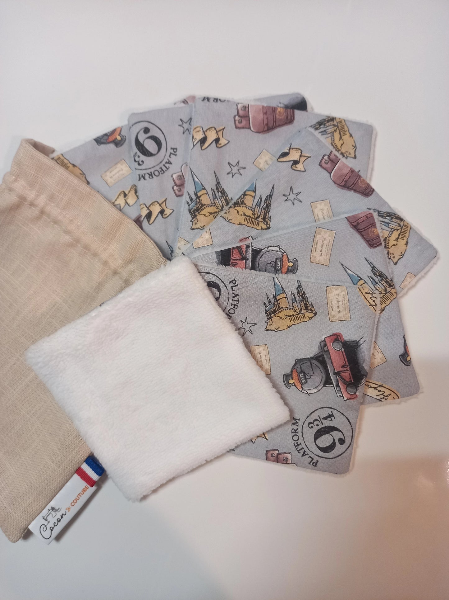 Lot de sept carrés démaquillants lavables au motif magique gris à train à vapeur, château, valises et étoiles, disposés en éventail à côté de leur pochon en lin Cocon Couture, un carré retourné sur le côté éponge blanc.