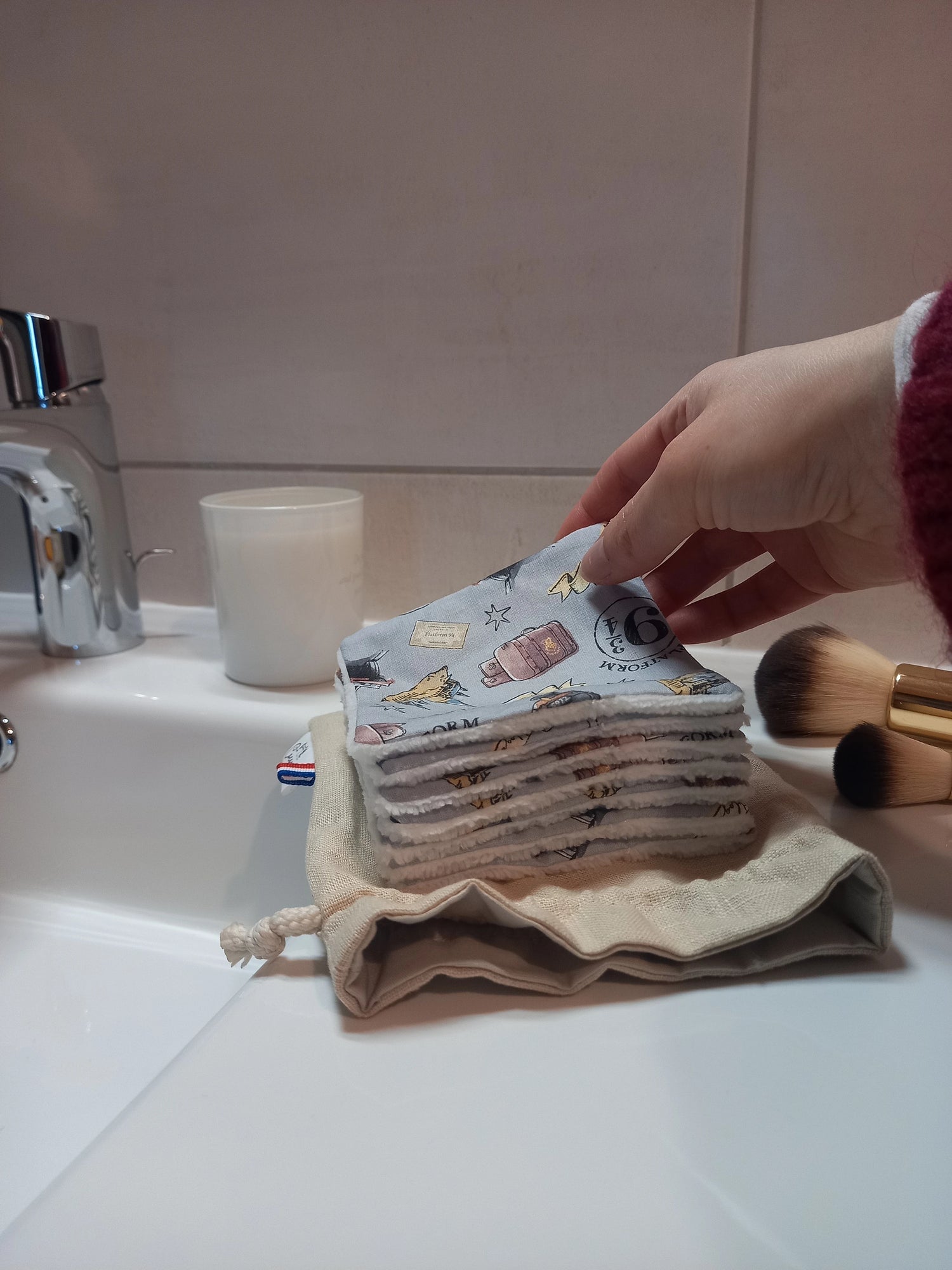 Carré démaquillant lavable au motif magique gris à train, château et valises tenu entre les doigts au-dessus de la pile posée sur le pochon en lin, sur le rebord d'un lavabo avec des pinceaux de maquillage.