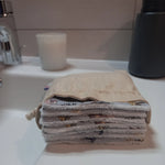 Carrés démaquillants lavables empilés dans leur pochon en lin posé sur le rebord d'un lavabo, le motif magique gris à train, château et valises ainsi que le côté éponge blanc visibles sur la tranche.