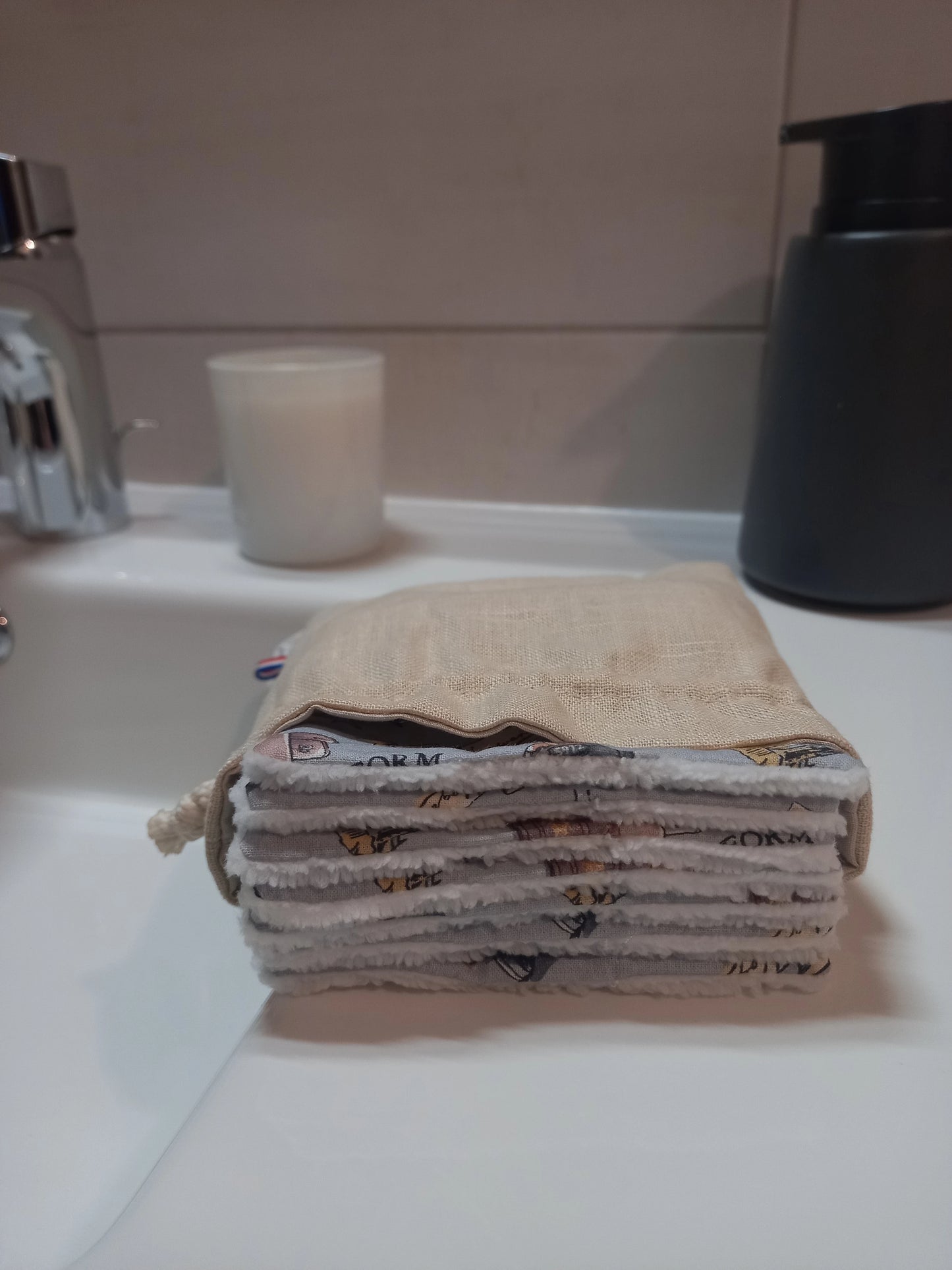 Carrés démaquillants lavables empilés dans leur pochon en lin posé sur le rebord d'un lavabo, le motif magique gris à train, château et valises ainsi que le côté éponge blanc visibles sur la tranche.