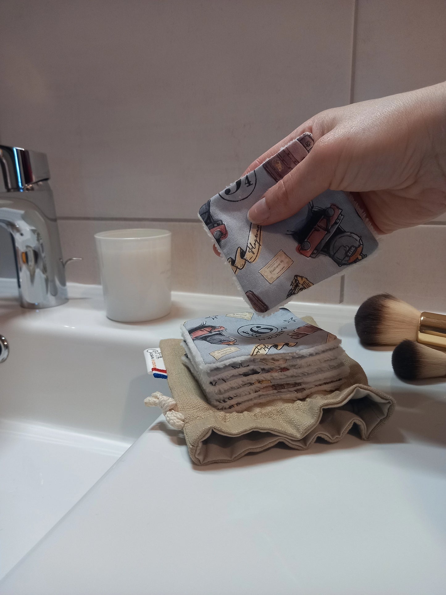 Main tenant un carré démaquillant lavable au motif magique gris à train, château et valises au-dessus d'une pile de carrés posée sur le pochon en lin, dans une salle de bain avec des pinceaux de maquillage.