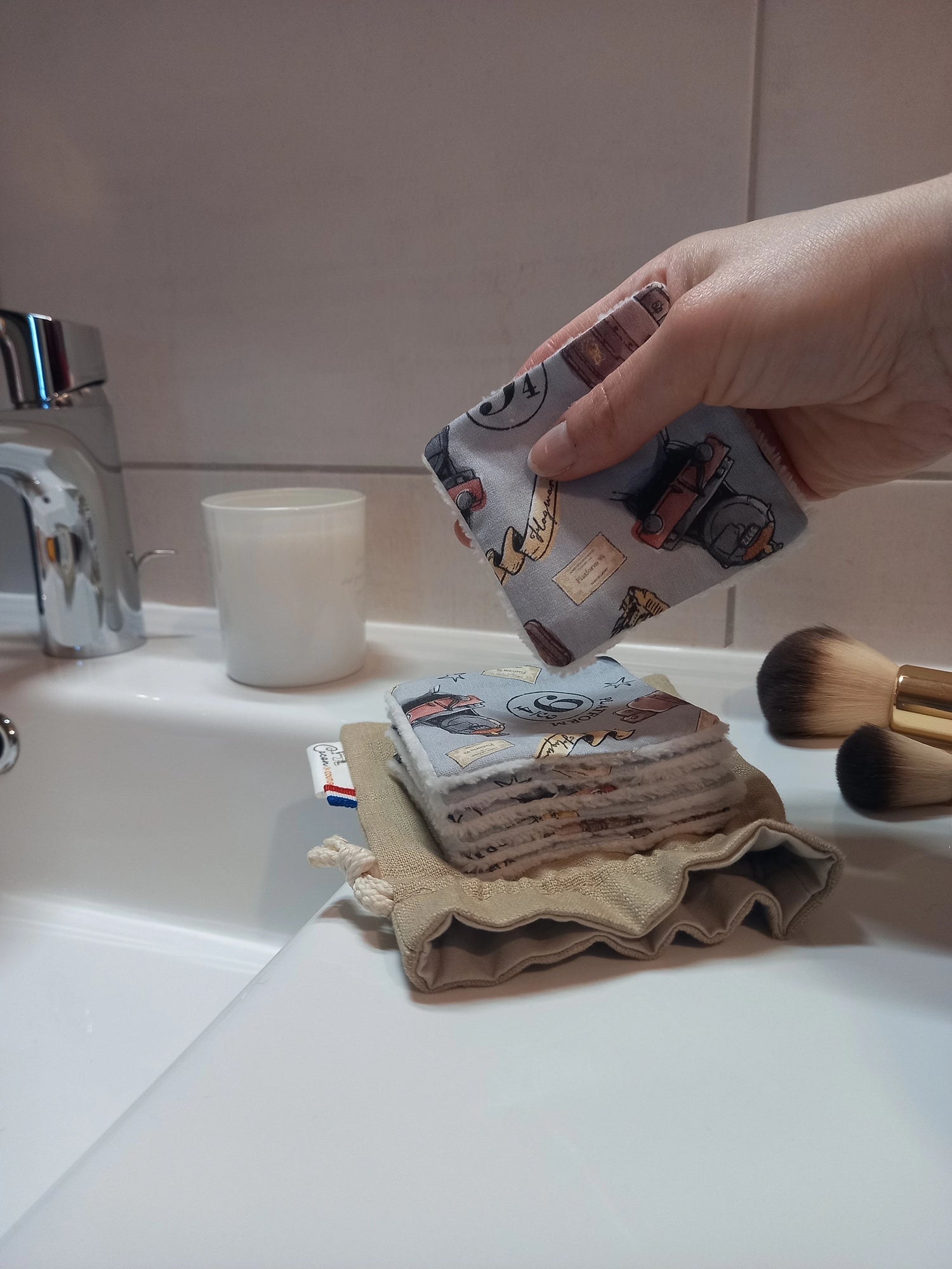 Main tenant un carré démaquillant lavable au motif magique gris à train, château et valises au-dessus d'une pile de carrés posée sur le pochon en lin, dans une salle de bain avec des pinceaux de maquillage.