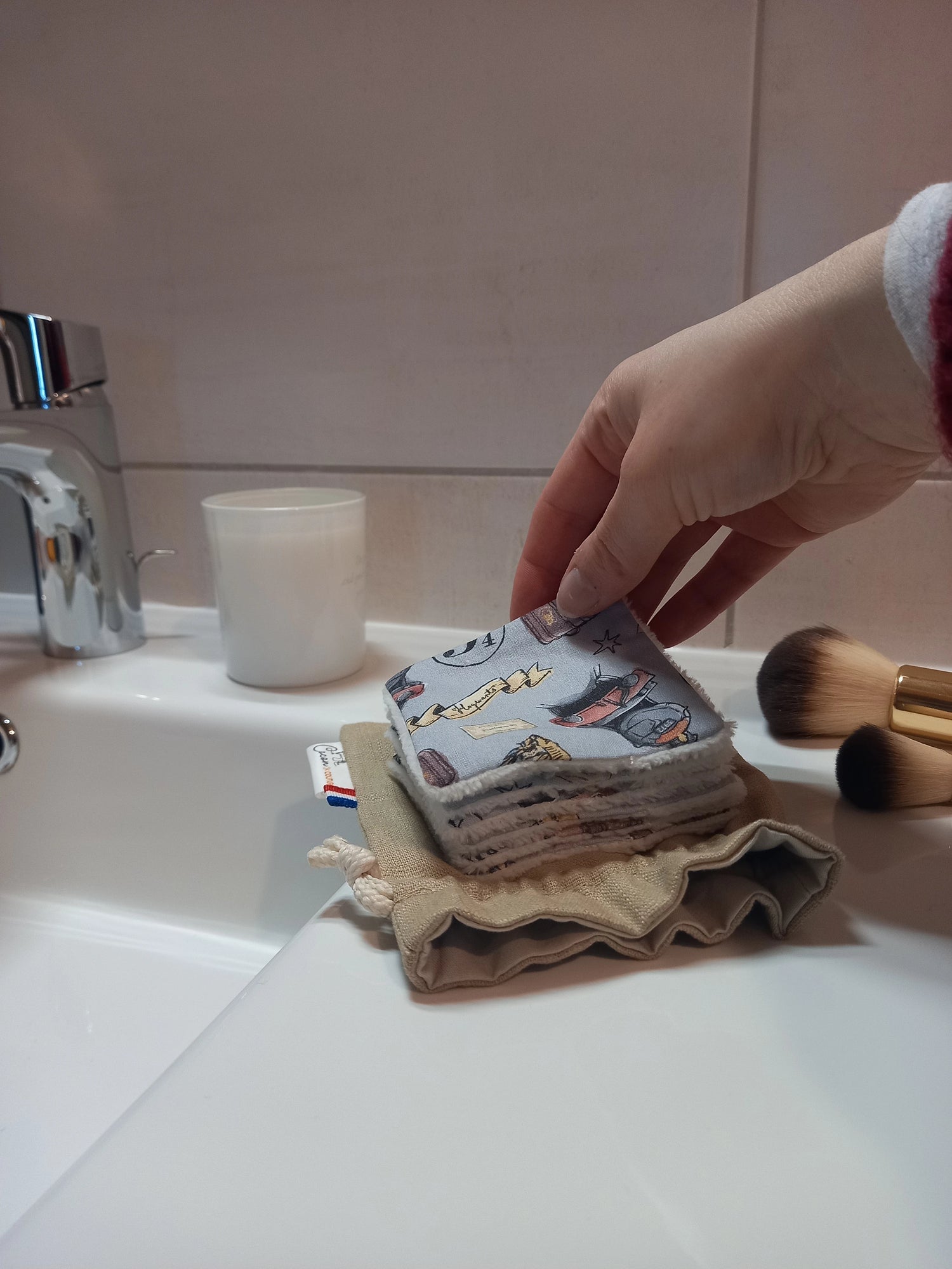 Carré démaquillant lavable au motif magique gris à train, château et valises tenu entre les doigts au-dessus de la pile posée sur le pochon en lin, sur le rebord d'un lavabo avec des pinceaux de maquillage.