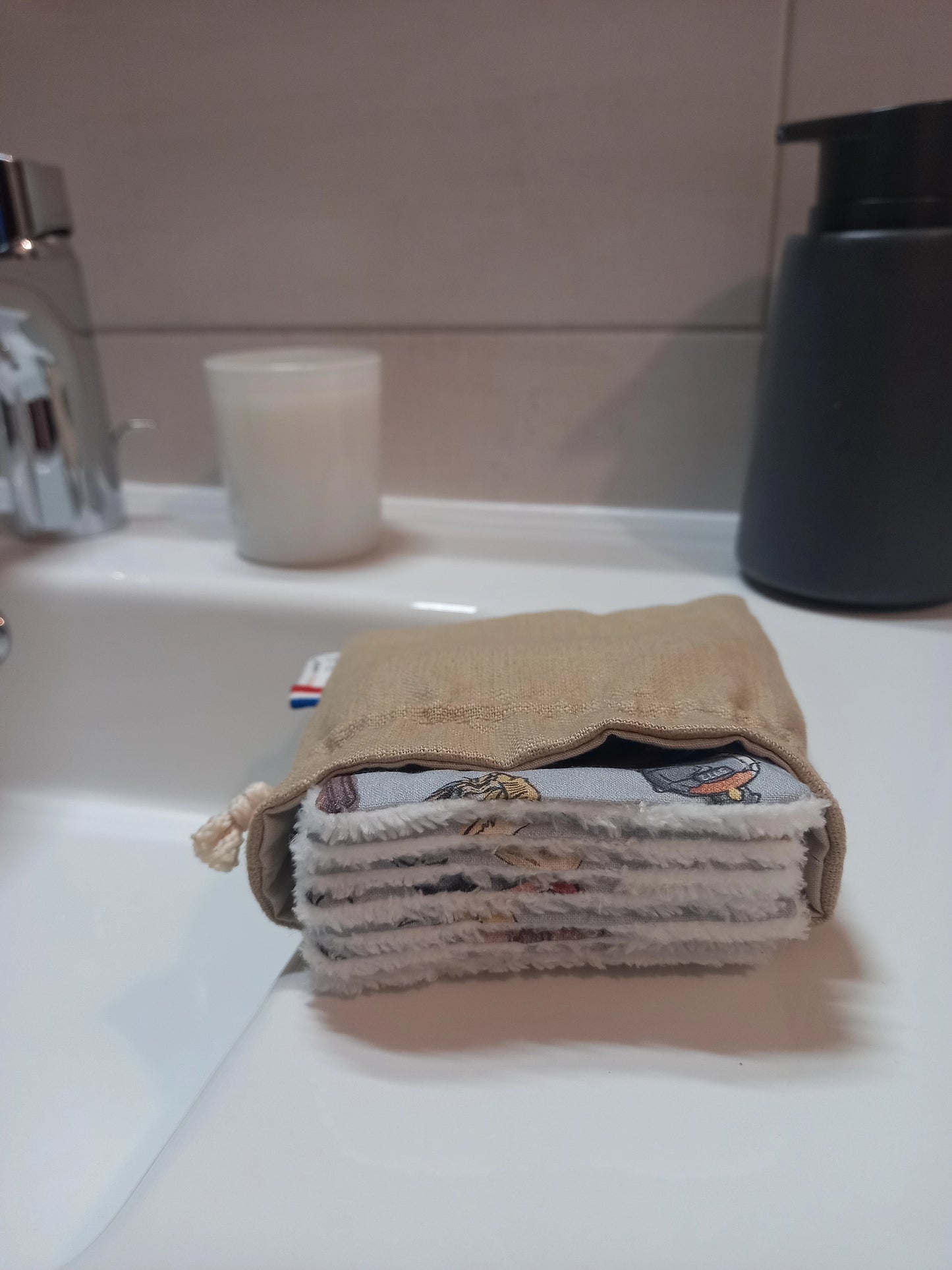 Carrés démaquillants lavables empilés dans leur pochon en lin posé sur le rebord d'un lavabo, le motif magique gris à train, château et valises ainsi que le côté éponge blanc visibles sur la tranche.