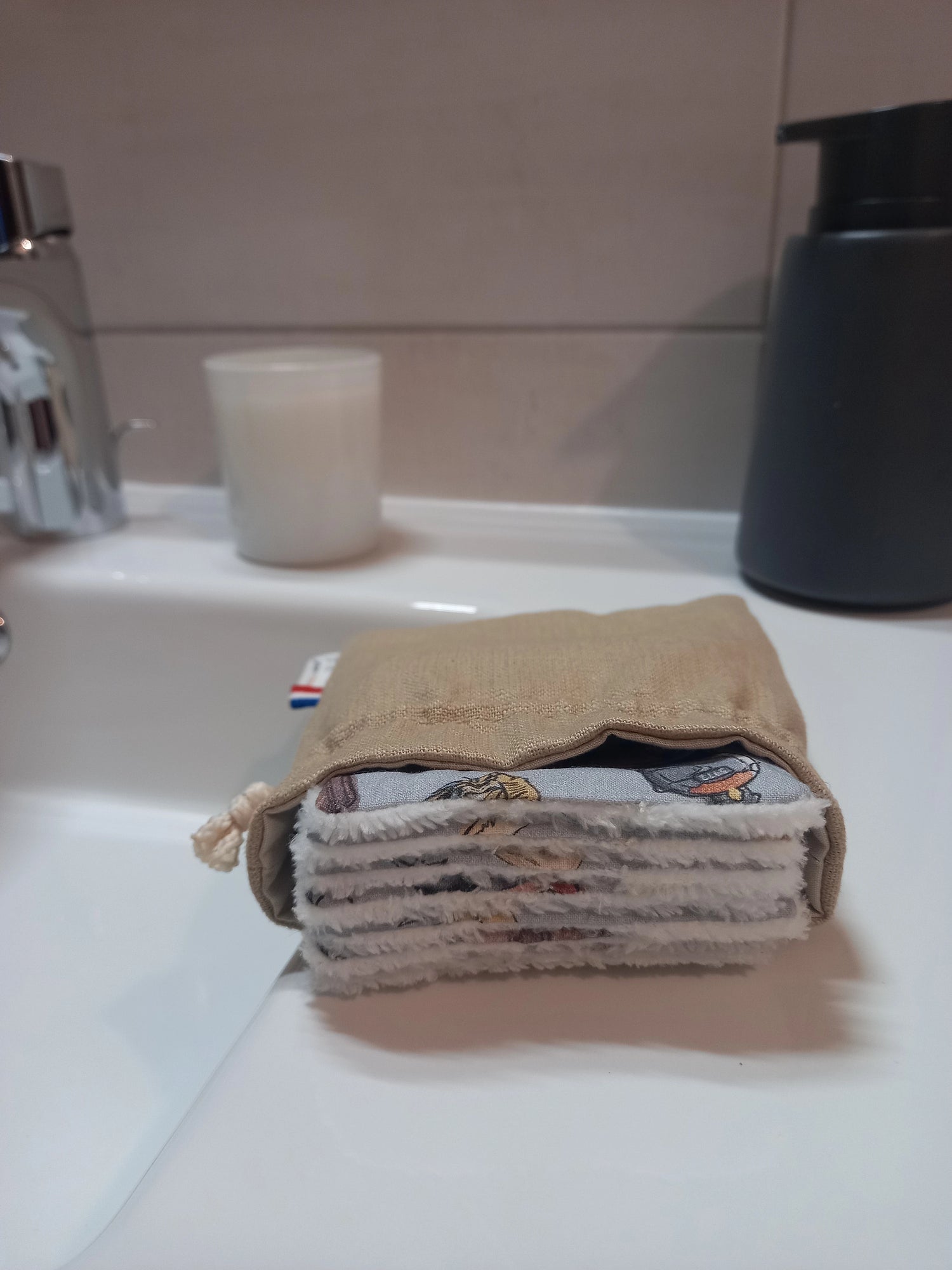 Carrés démaquillants lavables empilés dans leur pochon en lin posé sur le rebord d'un lavabo, le motif magique gris à train, château et valises ainsi que le côté éponge blanc visibles sur la tranche.