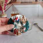 Charlotte alimentaire lavable à feuilles de palmier tenue à la main et retournée pour montrer son élastique froncé et son film alimentaire intérieur, avec l'étiquette Cocon Couture et un ruban tricolore, près d'un ramequin en verre vide.