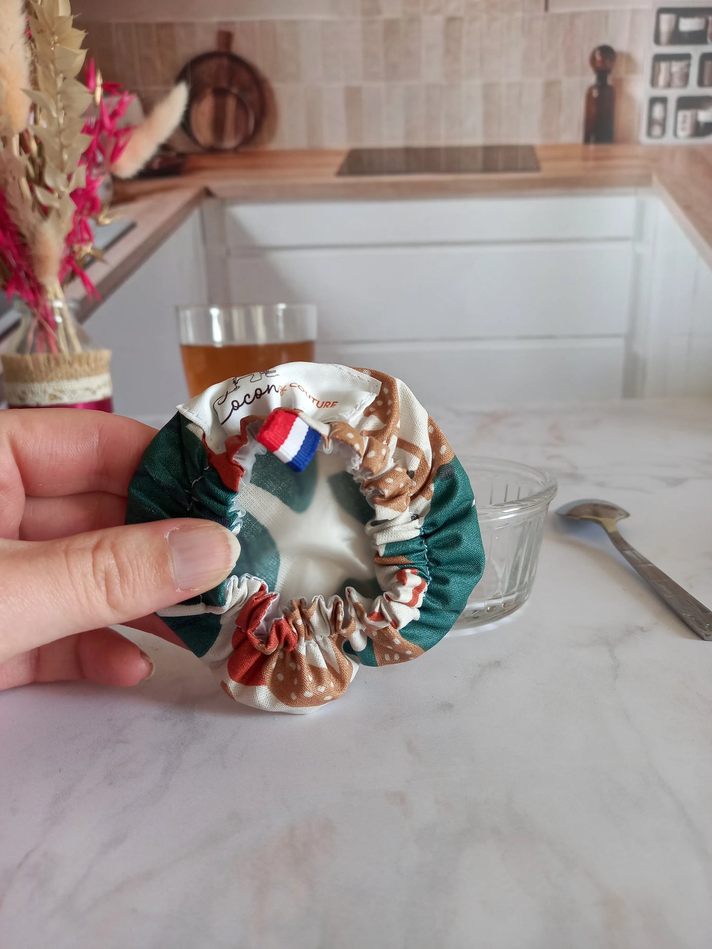 Charlotte alimentaire lavable à feuilles de palmier tenue à la main et retournée pour montrer son élastique froncé et son film alimentaire intérieur, avec l'étiquette Cocon Couture et un ruban tricolore, près d'un ramequin en verre vide.